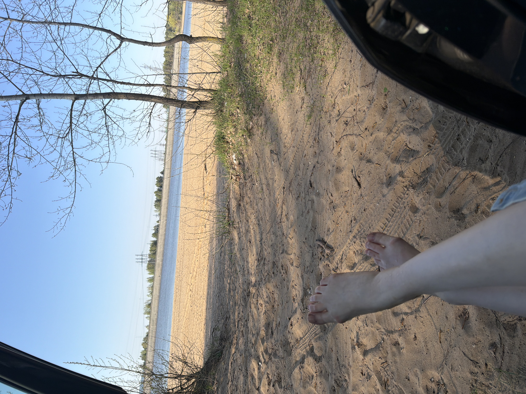 The image shows a beach scene, likely taken from inside a vehicle. The viewpoint is from inside a car, looking out towards a sandy beach. In the right foreground, two legs are crossed at the knees and appear to be pale skin, and a part of the car frame is visible. The sandy ground is littered with debris and tire tracks. The background depicts a shoreline, a body of water and power lines. Given the description of a beach in Noviy, Russia, it is likely the Volga River is visible. The sky is light blue and mostly clear, with bare trees and sparse vegetation in the foreground.