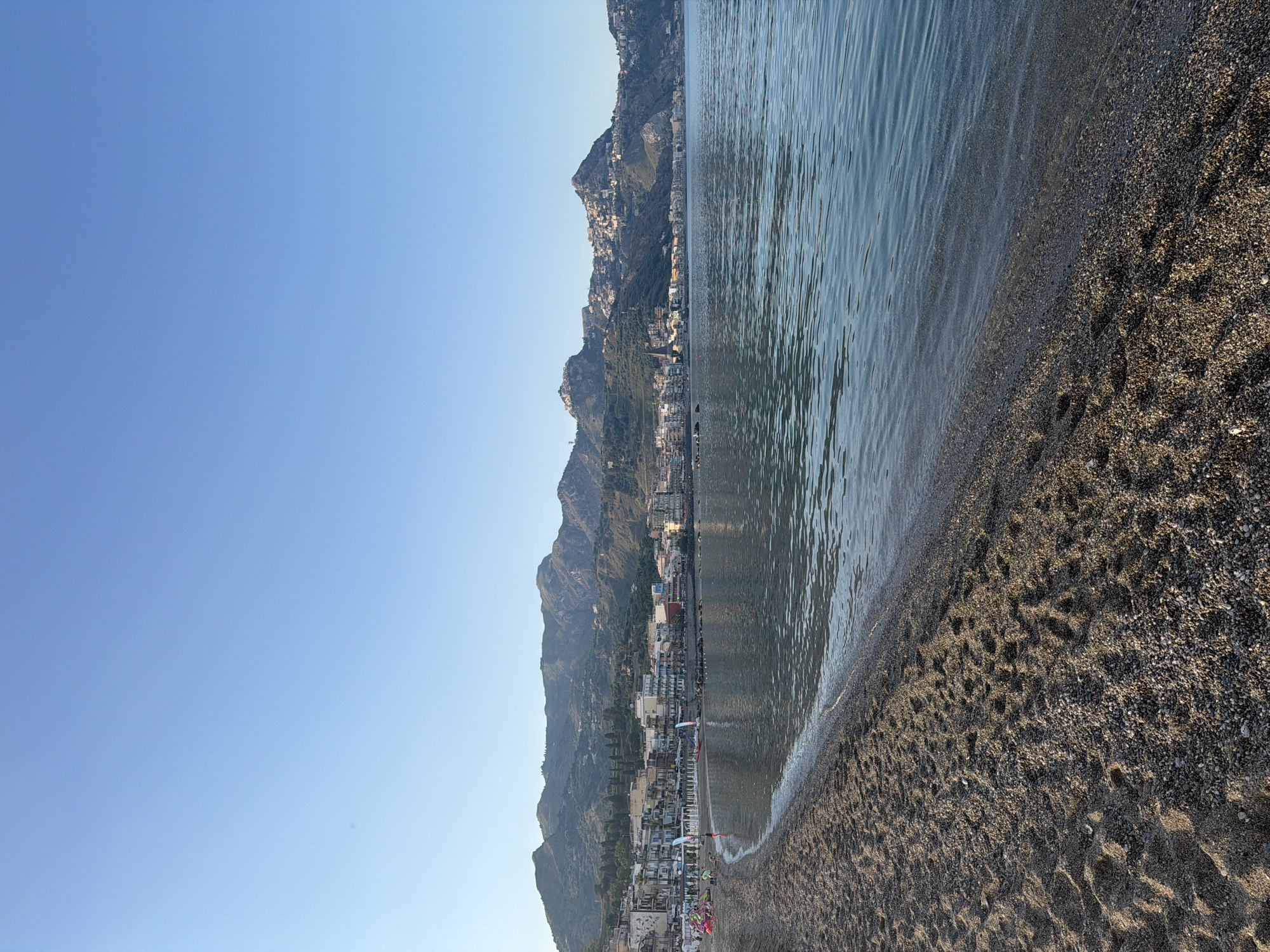The image shows a coastal view in Chianchitta, Italy. A beach with dark sand or pebbles occupies the foreground, transitioning to the water. A town with buildings nestled on a mountainside extends along the coast. The sky is clear and blue. There are a few people and umbrellas visible on the beach.
