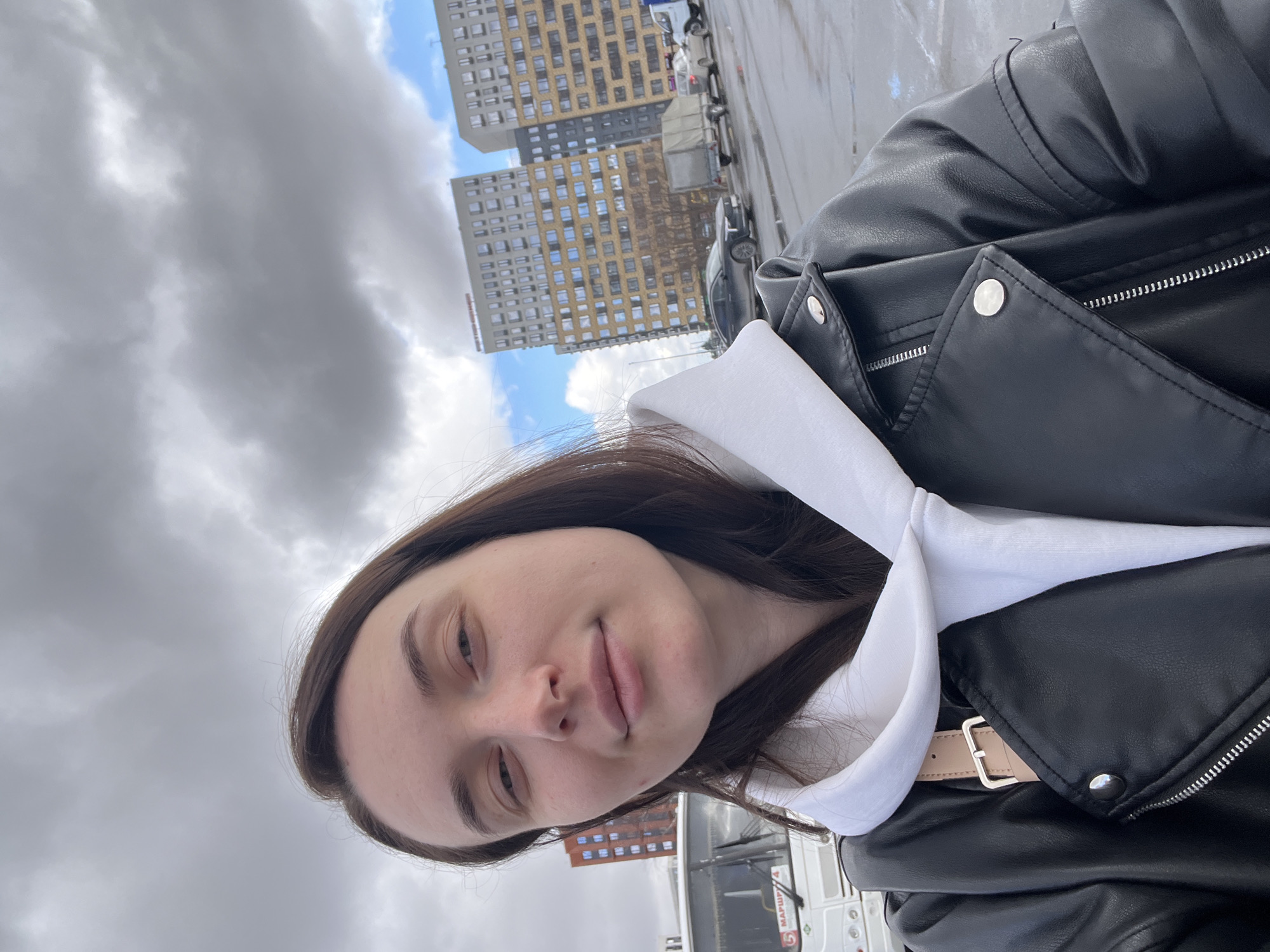 A young woman is taking a selfie outdoors. She is wearing a black leather jacket over a white hoodie. Behind her are buildings, cars, and a cloudy sky. Based on visual evidence, specifically the bus with the number 5 and the writing "МАРШРУТ 5", and the architectural style of the buildings, the location appears to be in the city of Tyumen, Russia.