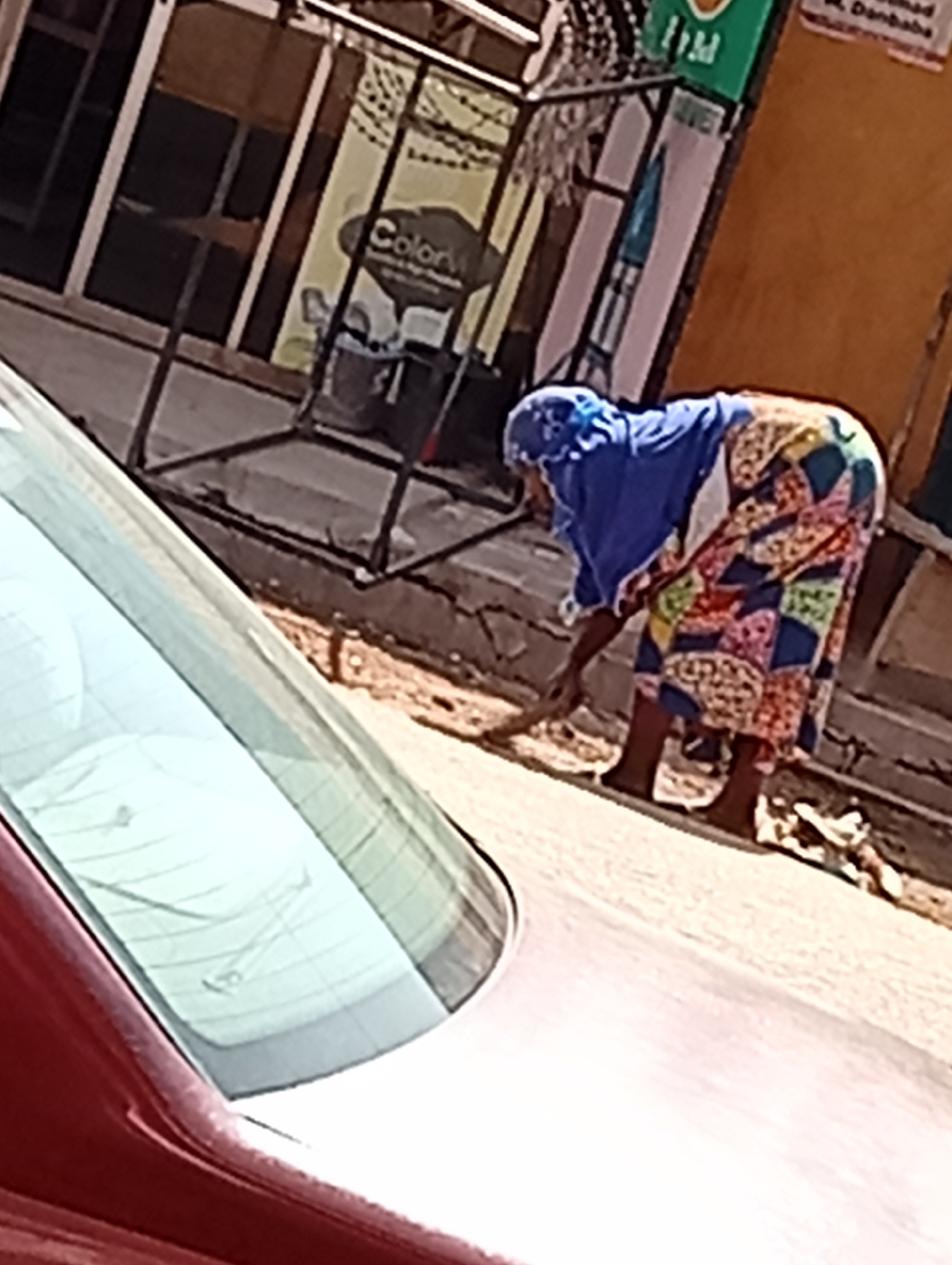 A woman wearing a blue headscarf and a long, colorful patchwork skirt is sweeping the street in front of a storefront. The location appears to be a street in Maiburiji, Nigeria. A red car is partially visible in the foreground. A sign with the word "Color" is seen on a store. A green sign labeled "PM" is also visible.