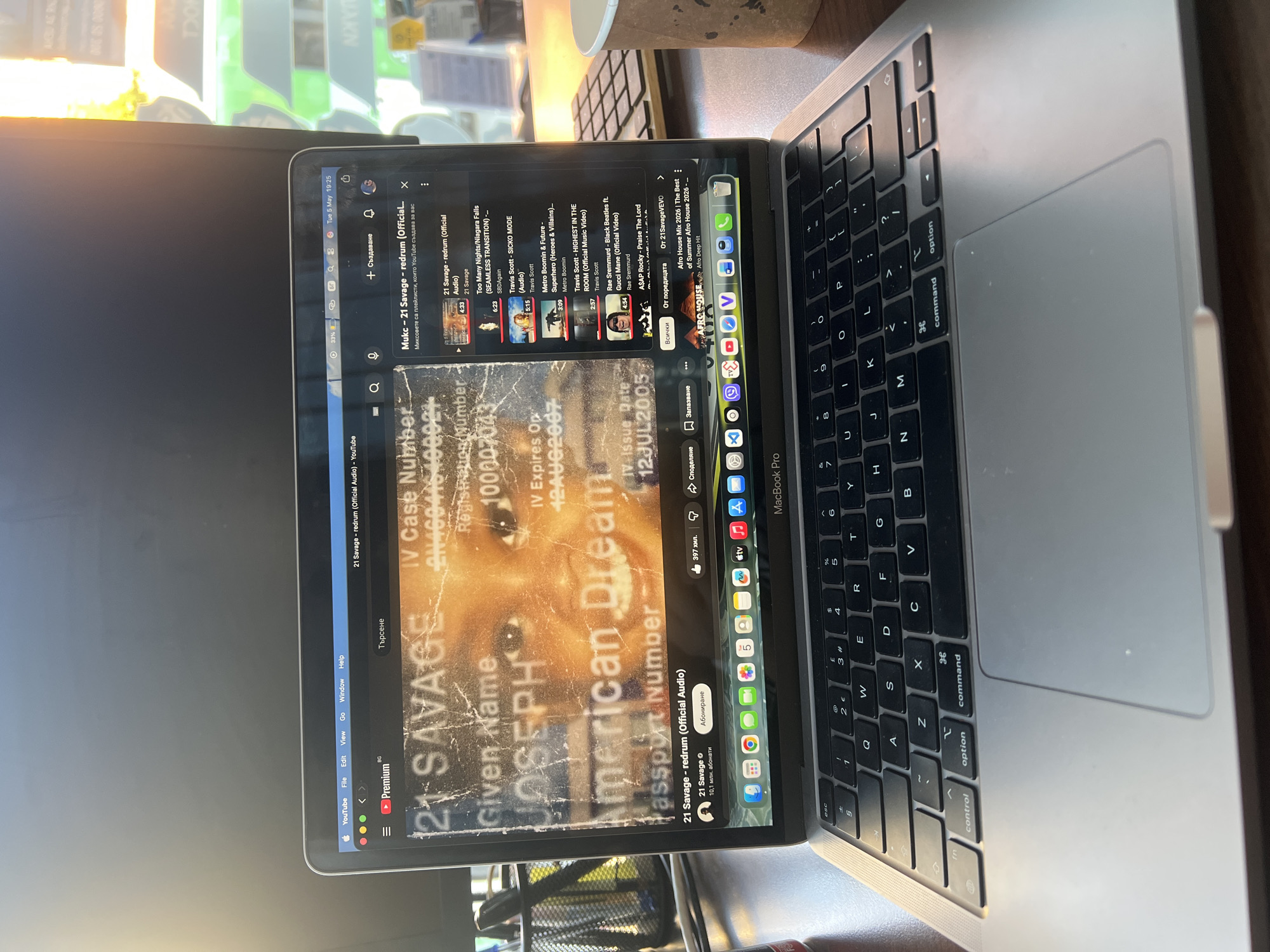 The image shows a MacBook Pro laptop displaying a YouTube page with the music video "21 Savage - redrum (Official Audio)". The screen also features an image of a passport or identification document with information such as "IV Case Number", "Registration Number", "Given Name Joseph", and "American Dream". The keyboard of the laptop is visible, as are surrounding objects such as a cup and a keyboard. The MacBook Pro appears to be open and positioned on a wooden desk or table. The image's contents include a search bar for content related to 21 Savage and other musical artists, suggesting a user browsing for music content. Based on the text in Cyrillic, the location is likely in Bulgaria, specifically Sofia.