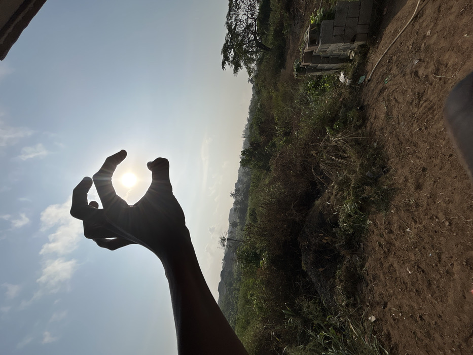 The image, reportedly captured in Debasco, Nigeria, displays an outdoor scene under a bright, partly cloudy sky. In the upper left quadrant, a silhouetted human left hand is positioned to partially frame and obscure the sun, which appears as a radiant disk with prominent light rays emanating from its circumference. The sky transitions from a light blue directly around the sun to a deeper blue further away, containing scattered white cumulus clouds.
Below the horizon line established by the hand, the foreground consists of uneven, reddish-brown earth mixed with sparse, dry vegetation and scattered debris. The mid-ground is dominated by an expanse of dense, uncultivated vegetation including various grasses, shrubs, and some smaller trees. Visible in the lower right, a stack of grey concrete blocks is present on a slight incline, alongside what appears to be a white plastic bag and a light-colored hose or pipe running along the ground. Further into the background, a large tree with a wide canopy stands on the right, and a hazy landscape featuring indistinct buildings and more greenery extends towards the horizon, indicating a semi-urban or developing area. No other individuals are visible in the scene.