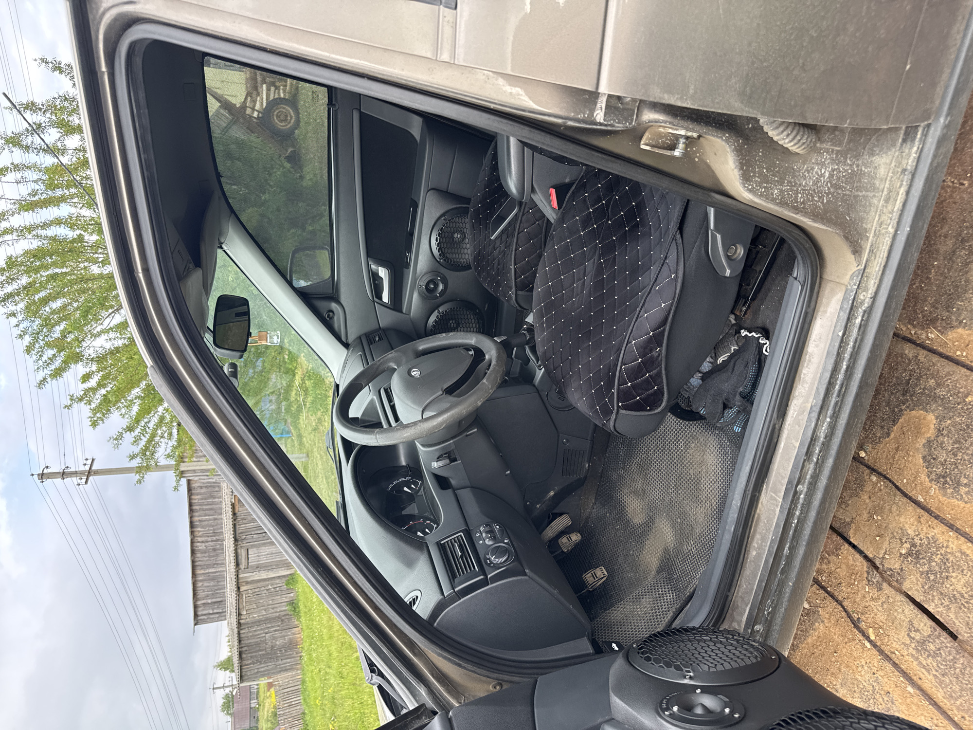The image displays the driver's side interior of a car with its door open, captured from an external viewpoint. The car is parked outdoors in Borodulino, Russia, under a bright daytime sky with scattered clouds.
Inside the vehicle, the steering wheel, dashboard, and a set of three pedals (clutch, brake, accelerator) are clearly visible. Both the driver's and partially the passenger's seats are fitted with distinctive black quilted covers adorned with white diamond-patterned stitching. The door panel features custom-installed speakers with mesh grilles, including two larger woofers and smaller tweeters positioned above them. The car's floor is covered with a dark, textured mat, which, along with the door sill and frame, shows noticeable dust and dirt, indicating recent use or an unpaved environment.
The background visible through the windows and open door shows a rural landscape. This includes green grass, trees, power lines strung between wooden poles, and a weathered wooden building or fence. A notable reflection of what appears to be an old tractor or similar agricultural machinery can be seen in the car's window. The overall impression is that of a functional vehicle in a rustic, countryside setting. No discernible text is visible in the image.