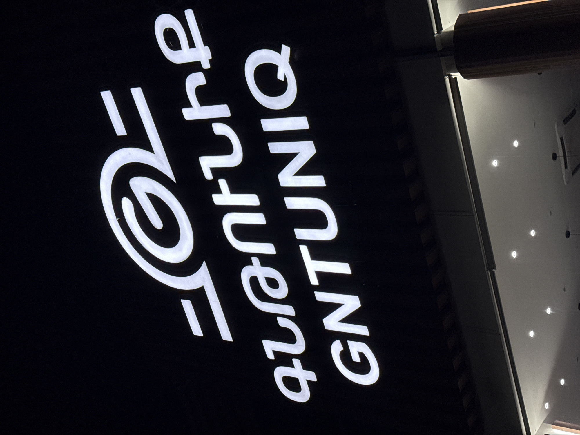 A nighttime shot showcases a sign reading "Future GNTUNIQ" illuminated in white. The sign is positioned on a dark background, and the letters are oriented vertically. Adjacent to the sign is a section of a building facade featuring recessed lighting. The location is Aparan, Armenia.