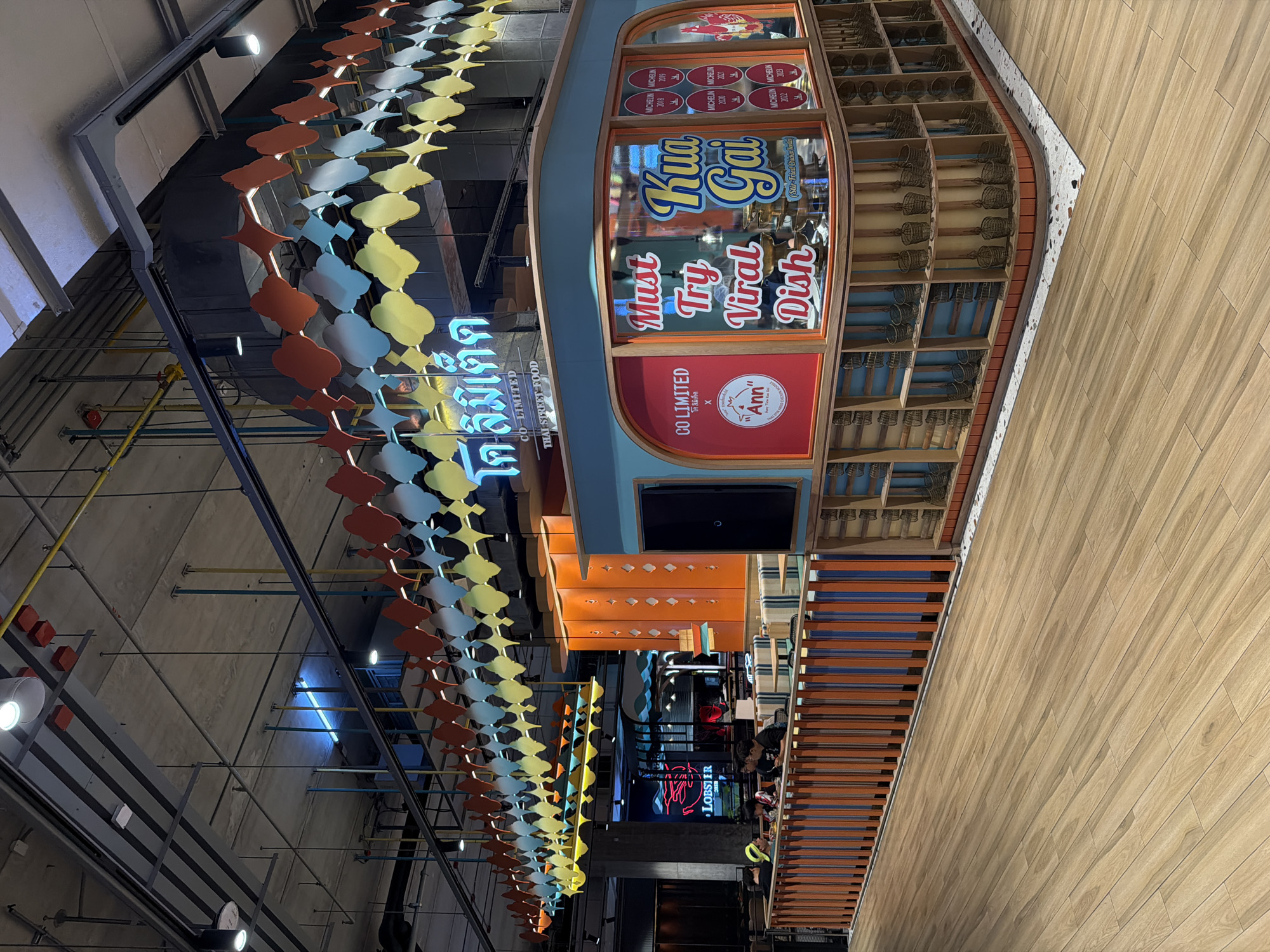 This image captures an indoor commercial space, likely a food court or market, featuring a vibrant food stall designed to resemble a boat. The stall is painted in shades of blue and orange, with prominent windows displaying text. Above the windows, several circular badges indicate "MICHELIN" awards for the years 2018, 2019, 2020, 2021, 2022, and 2023. The largest text on the window reads "Kua Gai" (with "Stir-Fried Chicken" in smaller print below) and "Must Try Viral Dish." Below the window, a red panel states "CO LIMITED x Ann," alongside a cartoon character. Inside the stall, shelves are filled with numerous traditional-looking woven baskets or cooking utensils.
To the left of the food stall, a long, colorful decorative structure hangs from the ceiling. It features alternating shapes resembling clouds or fish in yellow, light blue, and orange. Below this decor, a neon sign in Thai script reads "โค ลิมิเต็ด" (Co Limited) and "THAI STREET FOOD." Further in the background, another neon sign spells out "LOBSTER." The ceiling above is industrial-style, with exposed pipes, ducts, and various light fixtures, including spotlights and blue linear lights. The flooring or a large wall section on the right consists of light-colored wooden planks. A few people are visible in the background, particularly near the "LOBSTER" sign, but their activities are indistinct. The overall ambiance suggests a modern, lively dining environment, likely in Thailand given the Thai language and food references.