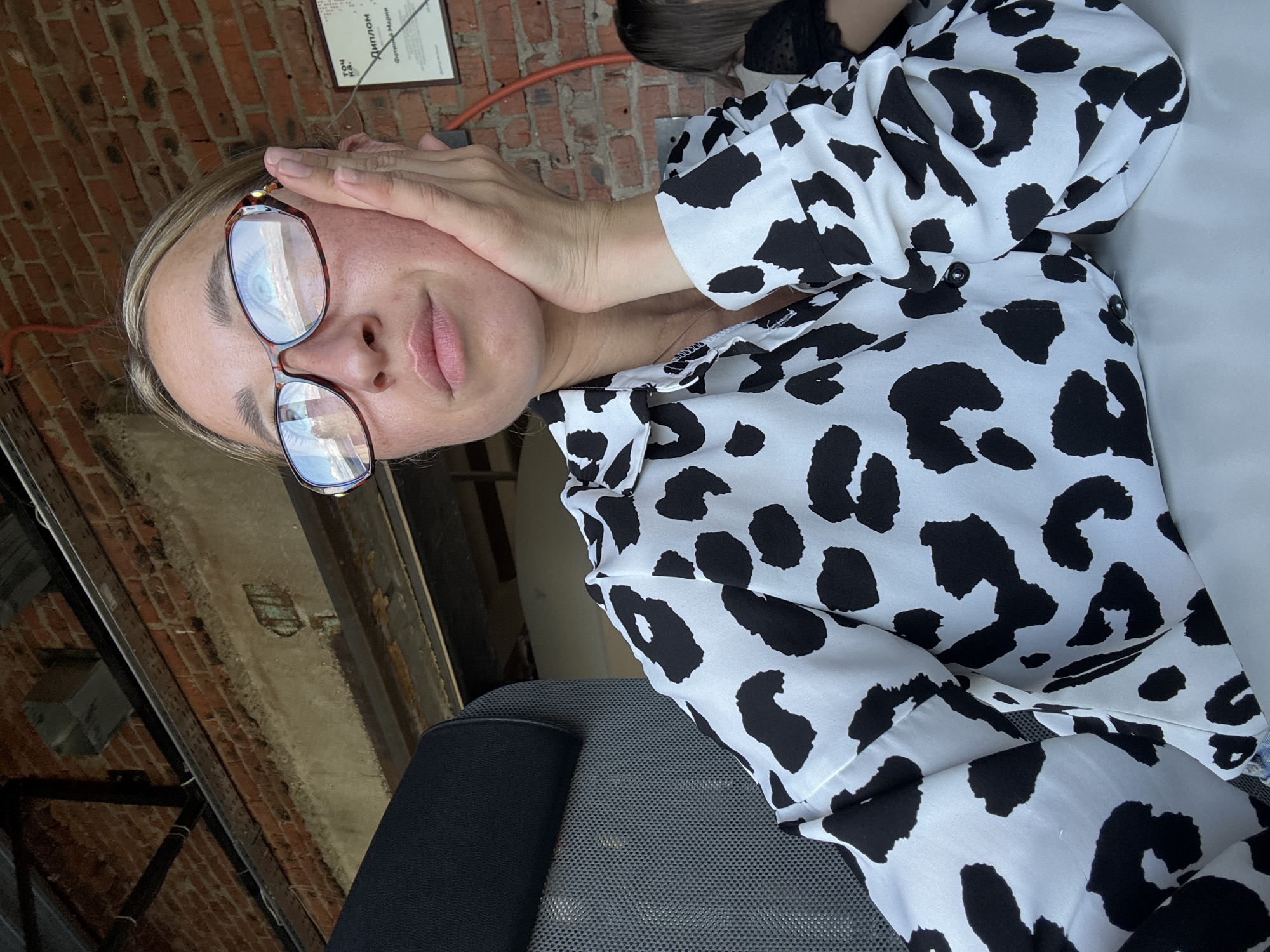 A woman with blonde hair and glasses is pictured sitting in a chair, resting her chin on her hand. She is wearing a white shirt with black leopard print. Behind her, a brick wall is visible, along with some electrical conduits and a framed document. A metal beam runs across the top of the frame, and what appears to be part of another person is visible to the right. The scene suggests an indoor location, possibly a cafe or office with an industrial aesthetic. The framed document is written in Russian and includes the word "ДИПЛОМ" (Diploma). This suggests the location could be Chaikovsky, Russia, given the language on the document.