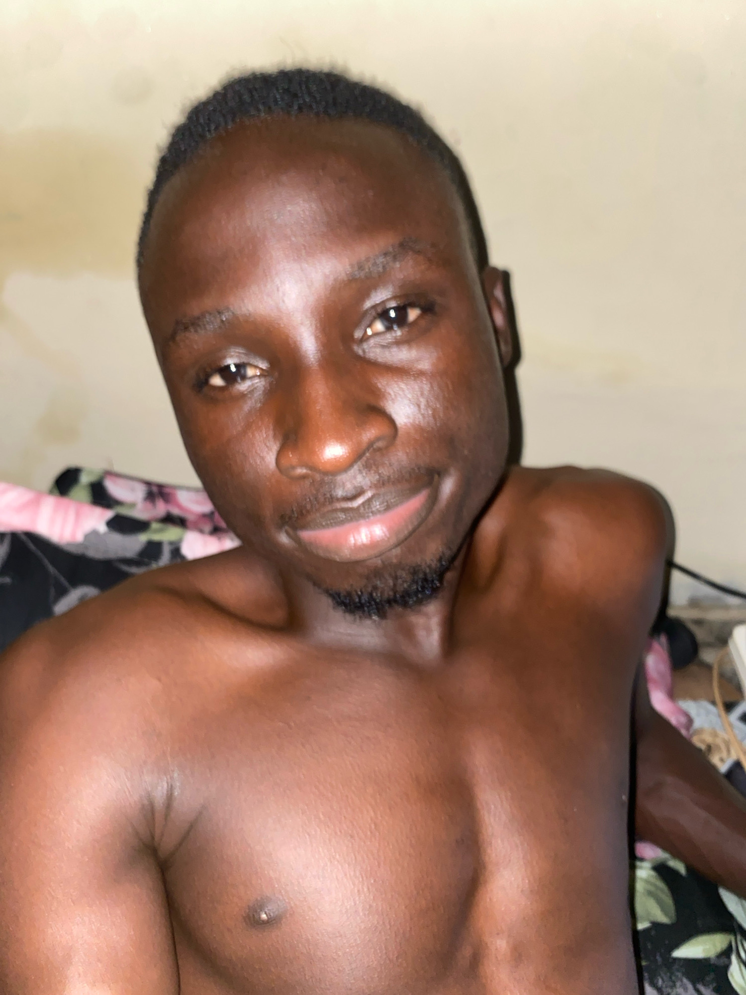 The image shows a close-up, waist-up shot of a young Black male with dark skin, wearing no shirt. He has short, dark hair and a small goatee. He is looking towards the camera with a slight smile. He is lying down, with part of a floral-patterned fabric visible behind him. The background is a plain, light-colored wall. The overall lighting is dim, suggesting an indoor setting, possibly a bedroom. There are no other individuals or obvious objects that would indicate a specific location within Barden Yana, Nigeria, beyond the general context of a domestic space.