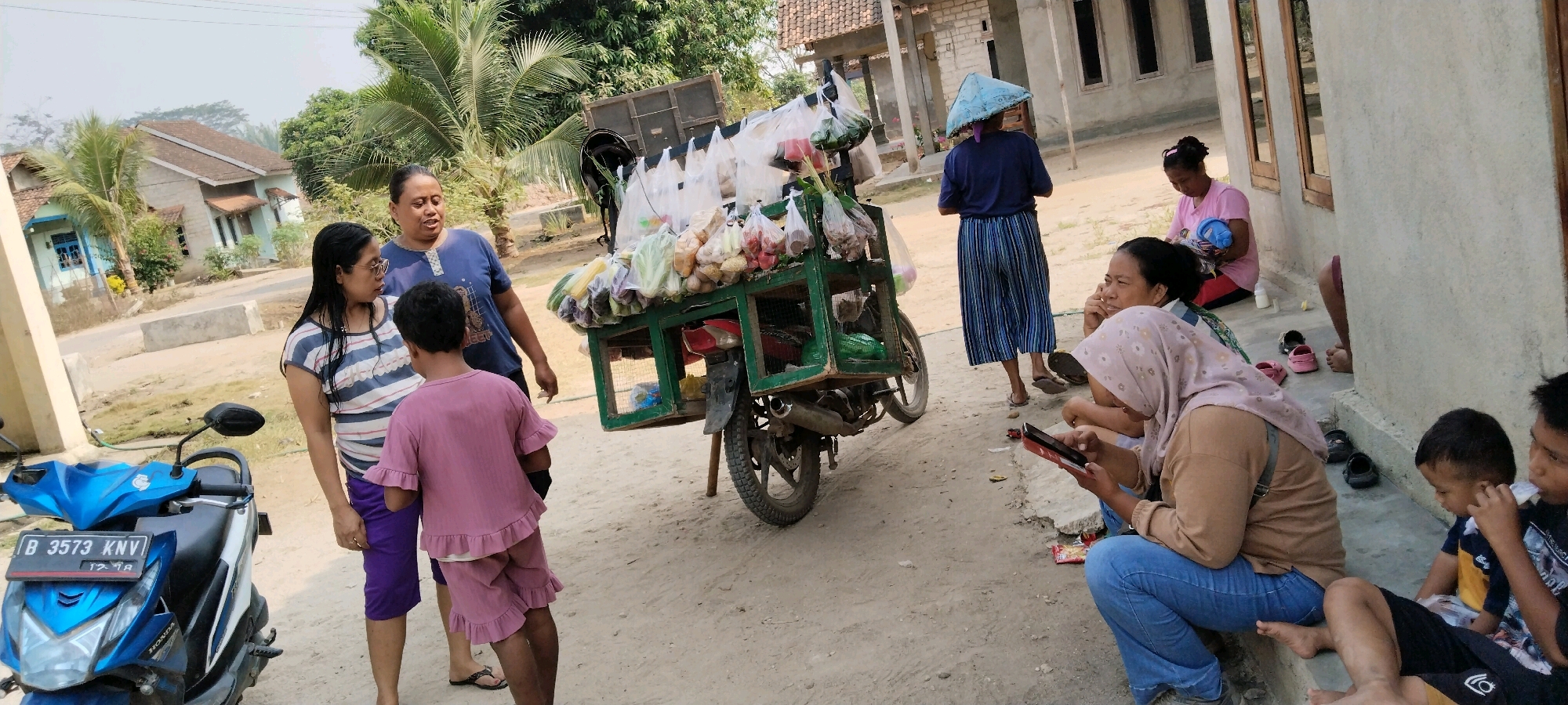 In Tanjung Ratu Ilir, Indonesia, on a dusty, sunlit day, a group of people are gathered near a house and a street. A motorcycle with the license plate "B 3573 KNV" is parked on the left. In the center, a two-wheeled cart, loaded with various fresh produce in plastic bags, is mounted on a motor vehicle, likely a motorcycle, which is partially visible. Several individuals are interacting around the cart.
On the left, a woman wearing a striped t-shirt and glasses is speaking with a young child dressed in a pink outfit with ruffled edges. Another woman, in a blue t-shirt, stands nearby. Further to the right, a woman wearing a conical hat and a striped skirt stands facing away from the camera. In the foreground on the right, two women are seated. One, wearing a headscarf and a tan top with blue jeans, is looking at a phone. Another woman, partially visible, appears to be talking. A young boy, wearing a blue and yellow shirt, is sitting on the ground to the right, with another person whose legs are visible. The environment suggests a residential area with a house visible in the background, featuring a tiled roof and wooden window frames. The overall scene depicts a casual outdoor gathering, possibly a market or a moment of social interaction on a street.