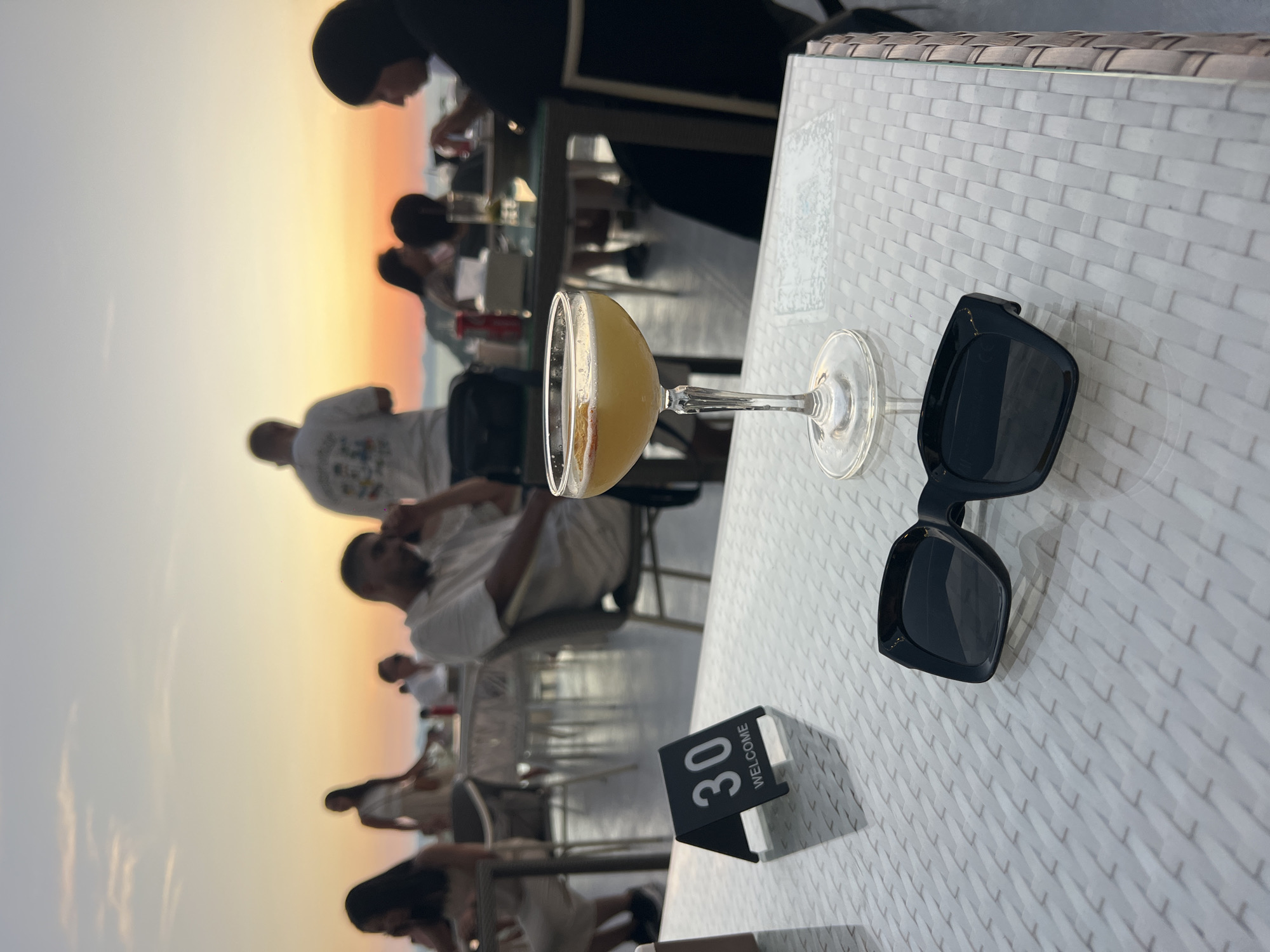 The image shows a table setting at a restaurant in Khao Thong, Thailand, at sunset. On the table are a coupe glass filled with a yellow beverage, a pair of black sunglasses, and a table number stand displaying "30" and "WELCOME." The table has a textured white surface. In the background, several people are seated at tables, presumably dining. The sky transitions from orange to light blue.