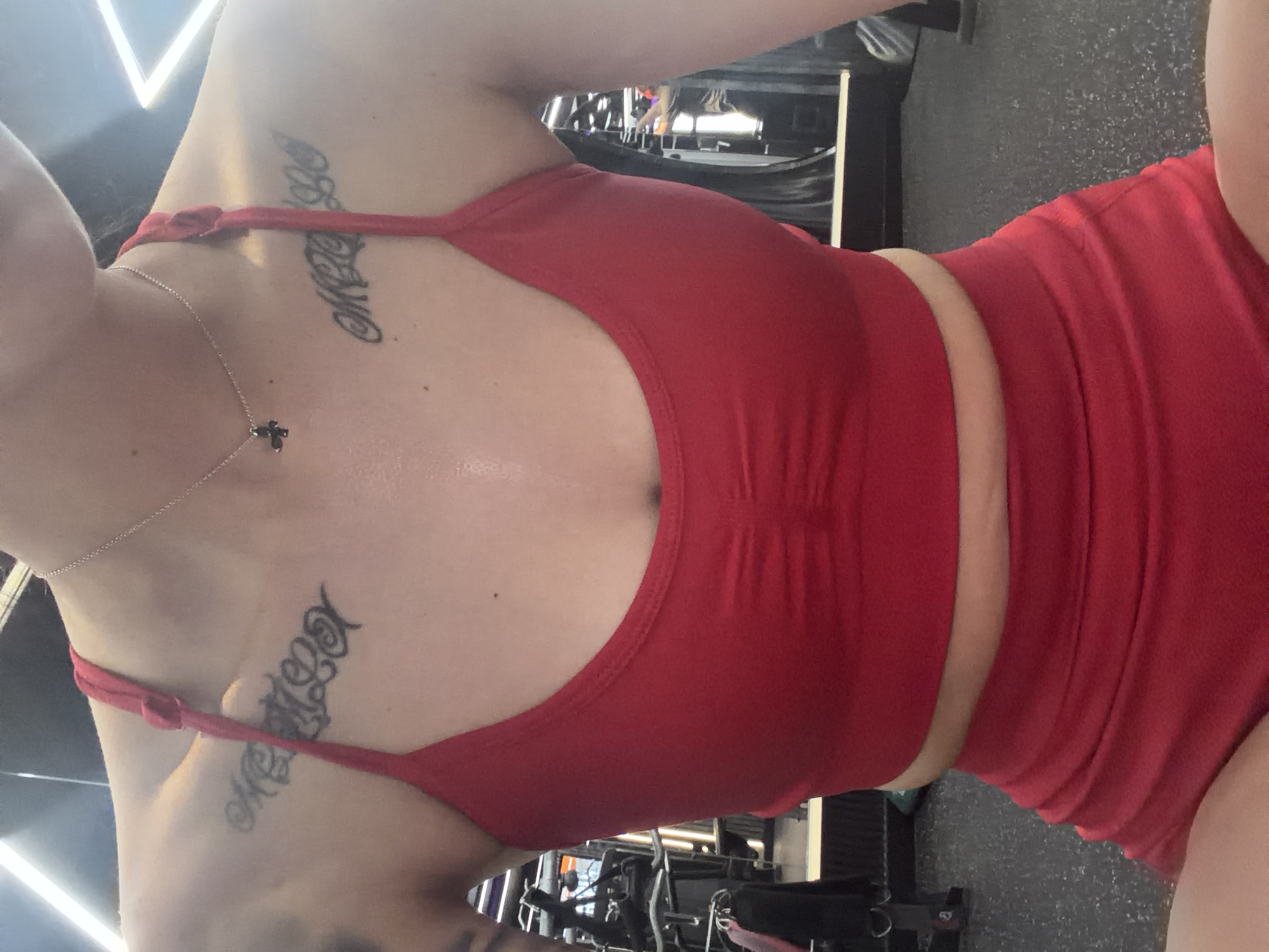 A person is wearing a red sports bra and shorts in a gym. The person has tattoos on their collarbone and chest area. The gym appears to be equipped with various weight machines and equipment. The lighting in the gym is bright, with fluorescent lights visible on the ceiling. The floor is a dark, textured material. The image is taken from a close-up perspective, focusing on the upper body of the person. No specific location in Plovdiv, Bulgaria is identifiable from the image.