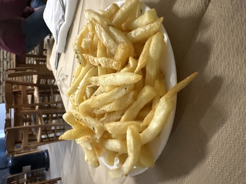 An overhead shot captures a large, overflowing oval white plate of golden-brown french fries, seasoned with dark green herbs (possibly oregano or parsley) and black specks (likely pepper). The fries are piled high, with some individual pieces extending off the edge of the plate.
The plate rests on a light brown or beige textured tablecloth. In the mid-ground, a white napkin and a metallic utensil are partially visible on the table. The background reveals an indoor setting, likely a restaurant, tavern, or cafe in Nea Erithrea, Greece. Numerous wooden chairs are arranged and stacked, suggesting a dining area. Behind the chairs, a wall constructed from light-colored stone or brick is visible, contributing to a rustic ambiance.
A person is partially visible in the far left background, seated at another table. They are wearing a dark top and blue pants, with their back turned towards the camera. A dark cylindrical object, possibly a waste bin or storage container, is at the bottom left, near the stacked chairs. The lighting appears to be artificial or ambient indoor light. No specific time of day, weather conditions, emotions, or visible text can be discerned from the image. The scene suggests a casual dining experience, with the fries presented as a side dish or appetizer.