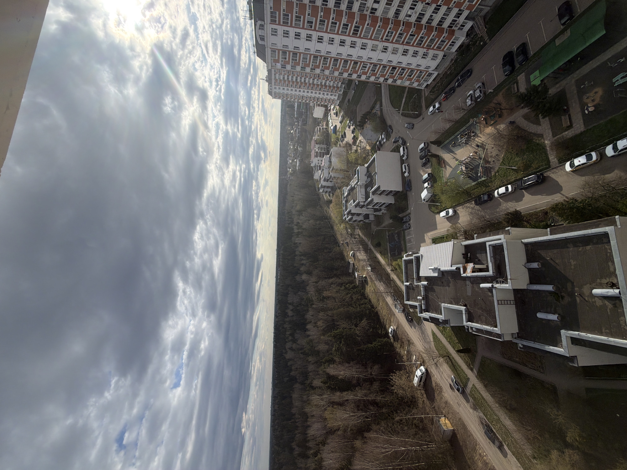 The image depicts an aerial view of a suburban area, likely in Brekhovo, Russia. The sky is predominantly cloudy with the sun partially obscured. On the right side of the image are several multi-story residential buildings, a mix of older, taller buildings with orange and white exteriors, and newer, shorter, white buildings with flat roofs. A road runs alongside these buildings, with parked cars and green spaces. A dense forest stretches along the center of the image, separating the built-up area from the skyline. Several cars are seen parked along the road that borders the forest. The scene is captured from a high vantage point, possibly from an upper floor of a building.