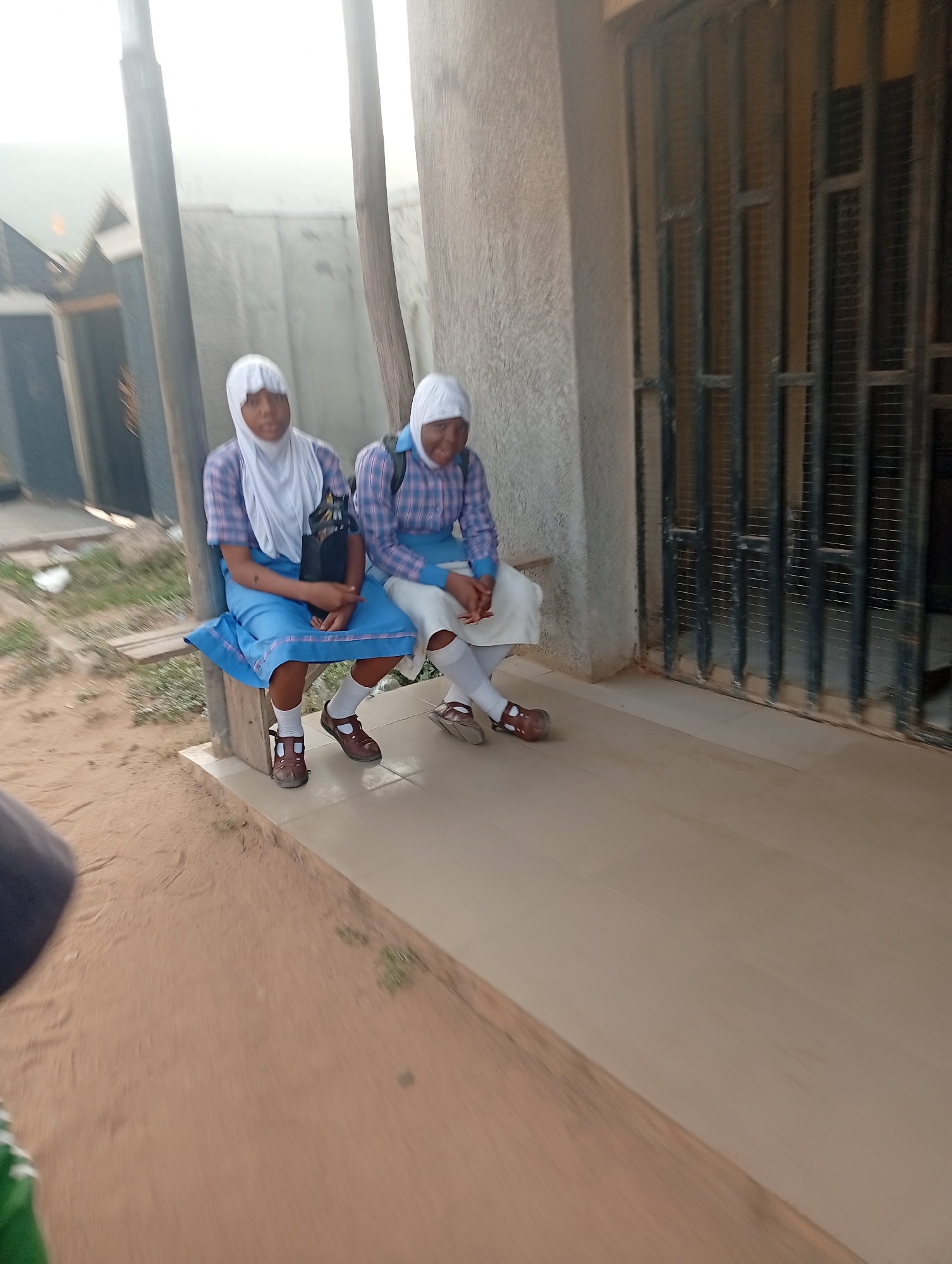 Two girls in school uniforms sit on a ledge next to a building. They wear white headscarves, plaid shirts, blue skirts, white socks, and brown shoes. One girl has a backpack. The location appears to be outdoors, possibly near a building with a barred window. The ground is a mix of dirt and concrete. This image was taken in the city of Lokoja, Nigeria.