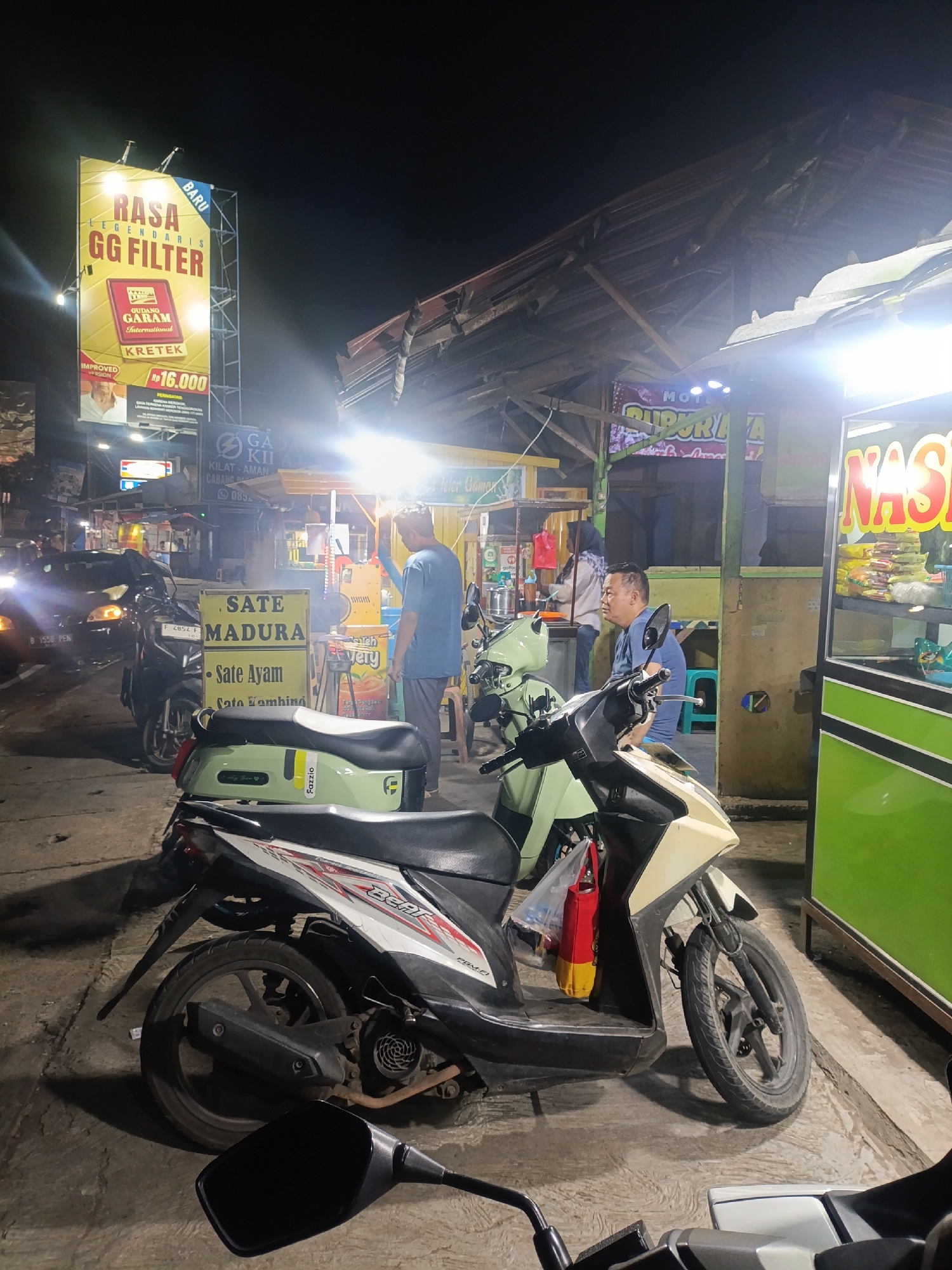 The image depicts a street scene at night in Jonggol, Indonesia. In the foreground, two motorcycles are parked. The one on the right is a light-colored scooter with a black seat and handlebars. The motorcycle on the left has a black and white design with red accents on the side. A large billboard advertising "RASA GG FILTER" and "KRETEK" is prominently displayed on the left side of the image. It also advertises Gudang Garam International for Rp 16.000.
Further back, several food stalls are visible. One stall has a sign that reads "SATE MADURA" and "Sate Ayam". Another stall displays "NASI". A man in a blue shirt is standing near the "SATE MADURA" stall, and another person is partially visible behind a stall on the left. A car is parked on the left side of the road, with its headlights on. The environment appears to be a typical Indonesian street at night, with overhead lighting and a rustic-looking roof structure over the stalls.