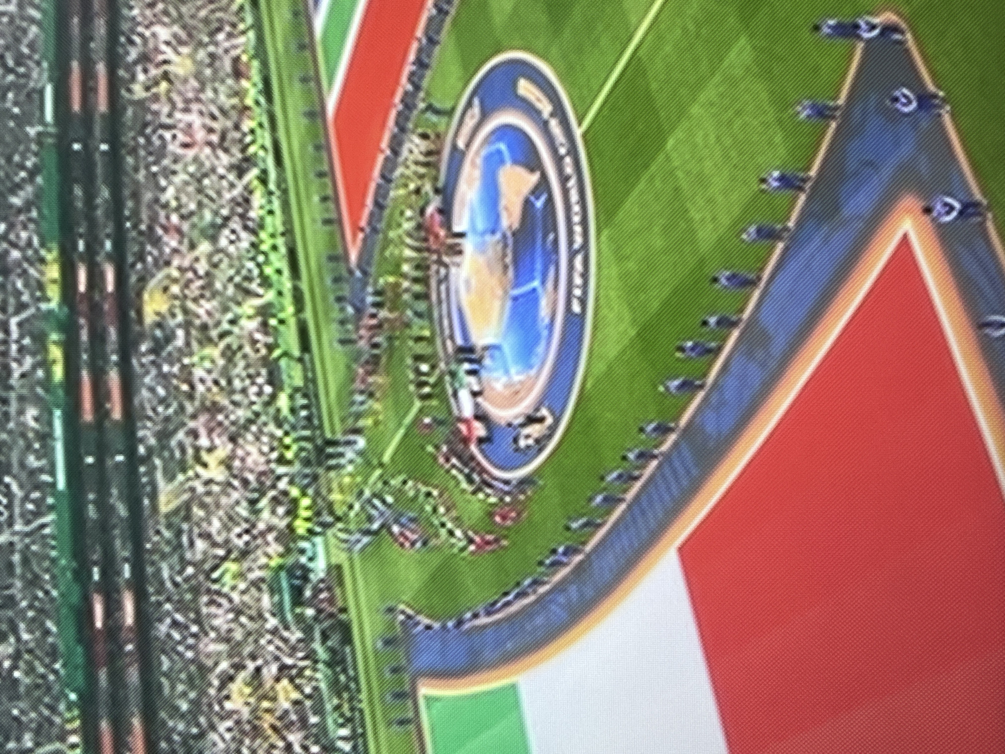 The image shows a football stadium with a large crowd in the stands. On the field, a ceremonial pathway is lined with individuals in blue uniforms. At the center of the field is a circular stage featuring a globe graphic with "FIFA WORLD CUP" written around it. Flags, one appearing to be the Italian flag, are visible on the field's edge.