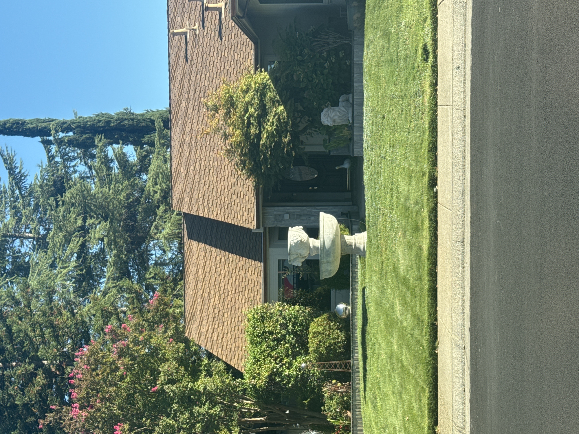 The image shows a residential house in Roseville, United States of America. The house has a brown tiled roof and is partially obscured by mature trees. In the front yard, a neatly mowed green lawn is visible, with a decorative fountain in the center. To the right of the fountain, a white lion statue is positioned in front of some landscaping. The house appears to be a single-story structure with an entrance visible in the center, flanked by windows and decorative elements. The environment suggests a sunny day with clear blue skies.