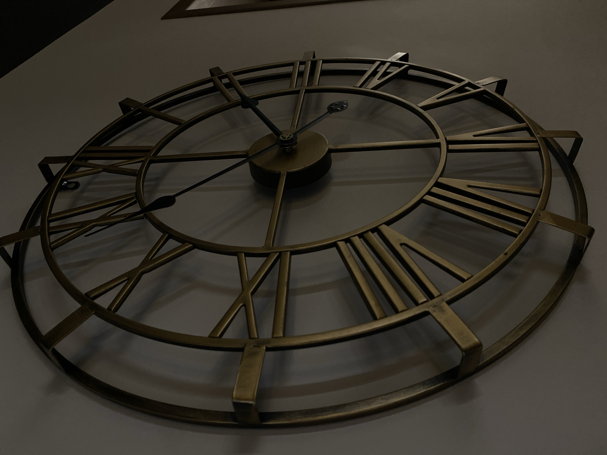 A low-angle shot shows a large, decorative clock hanging on a light gray wall. The clock has a bronze-colored metal frame and features Roman numerals to mark the hours. The clock hands indicate a time of approximately 1:52. The clock face appears to be open, revealing the wall behind the numerals and the inner workings of the clock. The lighting is dim, casting shadows across the wall and highlighting the metallic texture of the clock. The overall composition is a close-up view, focusing on the clock as the primary subject. There is no indication in the image of the city of Votkinsk, Russia.