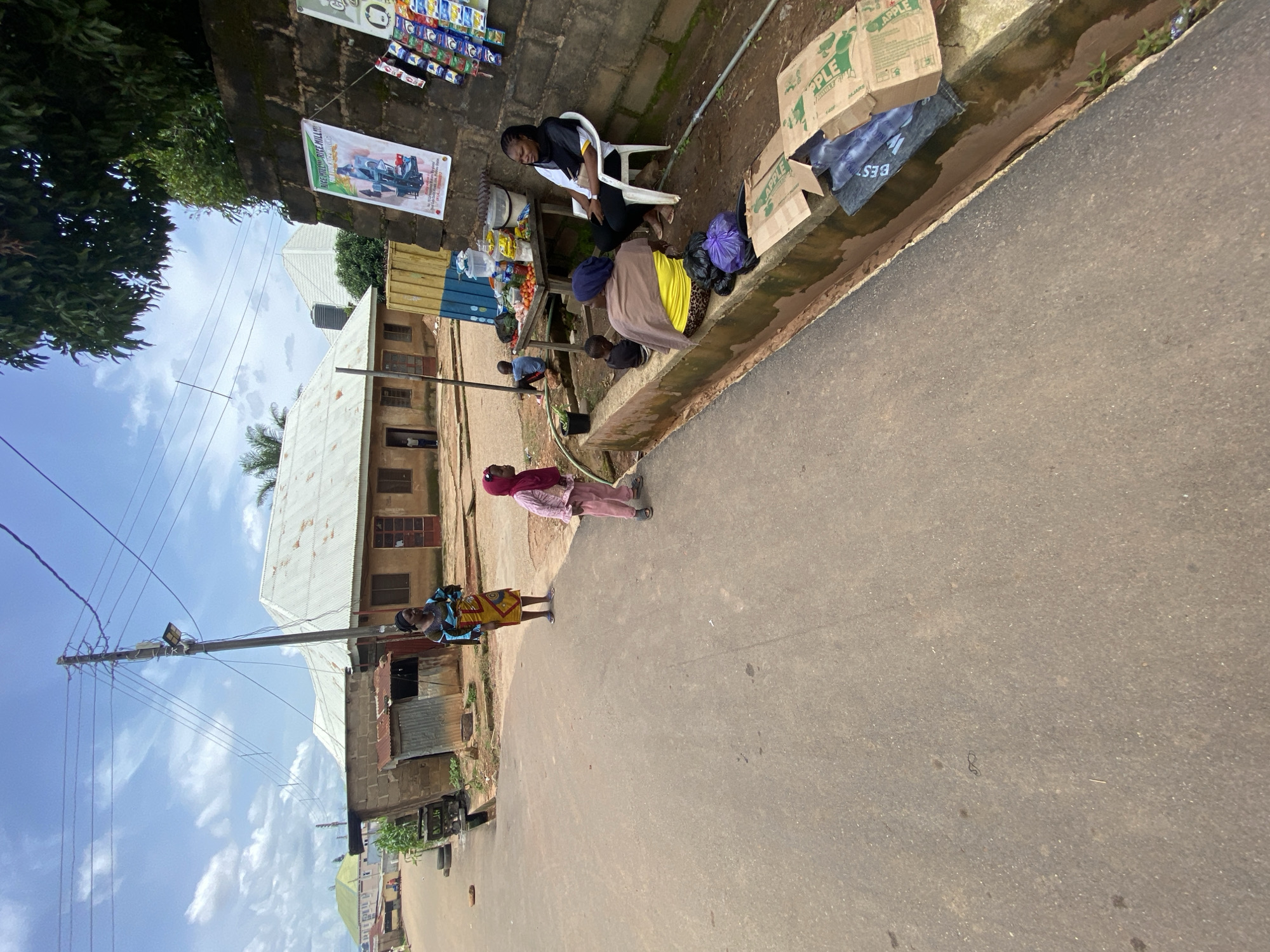 A street scene in Gura Top, Nigeria, shows a paved road with a drainage ditch along the right side. Several people are present: a woman in a patterned blue and yellow dress stands near a utility pole. A young girl in a pink outfit and headscarf stands on the road. Near the ditch, a woman in a yellow top and a headscarf sits with two small children. Another woman sits on a white chair by a stall displaying food items and various packaged goods. Several cardboard boxes and plastic bags are on the ground near the ditch. Buildings line the street, one with a corrugated metal roof and another with a lighter-colored roof. A poster advertising a rice mill is attached to a brick wall. Power lines crisscross the sky, and trees are visible.