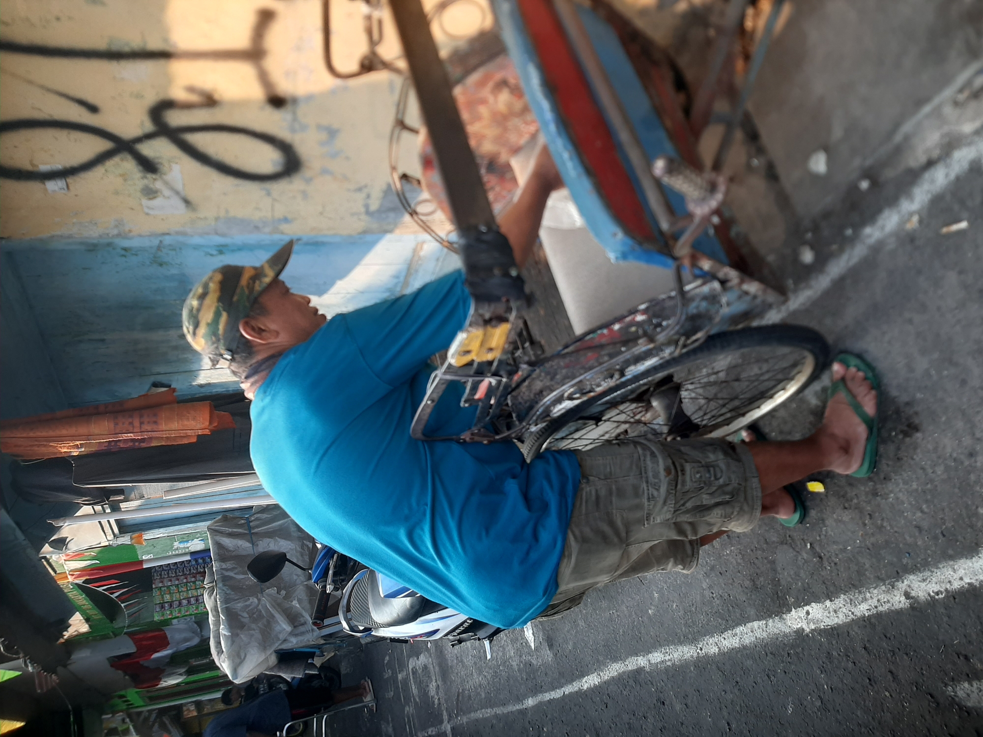 A middle-aged East Asian man wearing a camouflage baseball cap, a bright blue t-shirt, and tan cargo shorts stands next to a motorcycle and a three-wheeled rickshaw. The man is bent over, appearing to be adjusting something within the rickshaw. He wears green flip-flops. The rickshaw has a blue and red painted body, and a black metal frame. The motorcycle is white and blue. The scene appears to be a street in a market area, with stalls and buildings visible in the background. Clothes hang from an overhead line. The ground is asphalt, with white painted lines. Graffiti is visible on a weathered wall in the background. The lighting suggests it is either early morning or late afternoon. The location is identified as Central Jakarta, Indonesia.