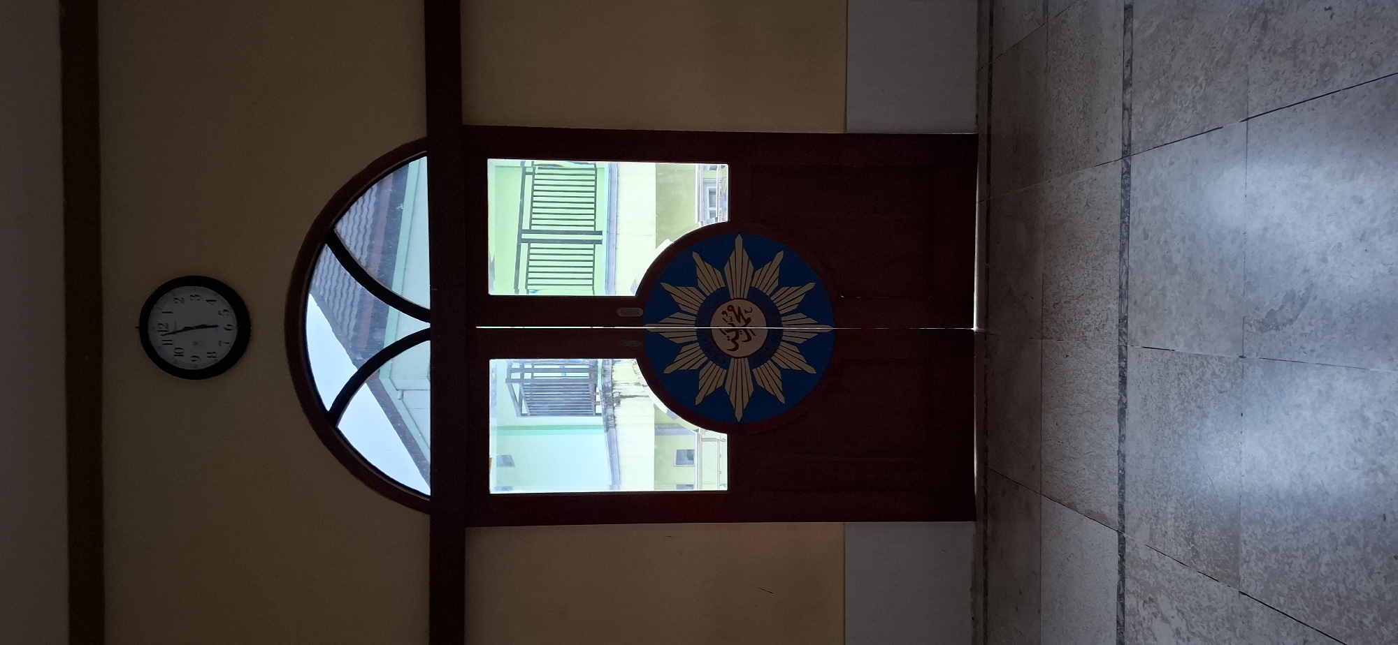 A digital photograph depicts a double wooden door with a round emblem in the center. The emblem features a blue circle with radiating yellow lines, and an Arabic inscription in the middle. Above the door is an arched window made of glass panes separated by dark wooden dividers. A black analog clock with white numbers and hands is mounted on the wall above the window. The wall is painted a light yellow, and the floor is tiled with large square marble-patterned tiles. In the background, through the glass panes of the door and the arched window, buildings with green and yellow facades and dark railings on balconies are visible. The scene appears to be indoors, looking out onto an outdoor environment. The image is lit with artificial light, casting shadows on the floor.