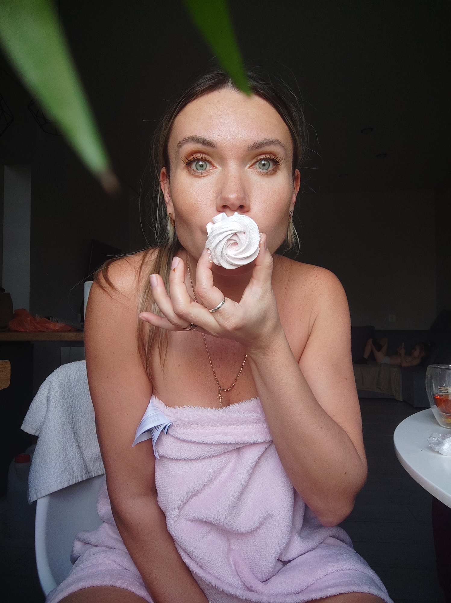 A woman with light brown hair and striking green/hazel eyes is pictured indoors in Votkinsk, Russia. She looks directly at the camera with a wide-eyed, expressive gaze, holding a swirled pink and white marshmallow or meringue to her mouth. She is wrapped in a fluffy pink towel, and another white towel is draped over the back of the white chair she is seated on. A gold necklace with a cross pendant hangs around her neck, and she wears a silver ring and another gold-toned ring on her left hand. Gold hoop earrings are visible. Her arms and shoulders show a scattering of freckles.
In the blurred foreground, large green plant leaves create soft, bright streaks across the upper left of the frame. The background is mostly dark, suggesting a dimly lit interior. On the right, a white table holds a clear double-walled glass containing a reddish-brown liquid, likely tea, with some indistinct items next to it. Further back, on a dark couch, a small child or person is vaguely visible lying down. To the left, a counter or shelf area shows a rumpled orange plastic bag. Natural light illuminates the woman from the front, highlighting her features. A white care label sticks out from the pink towel. Faint, unreadable text is partially visible on the glass.