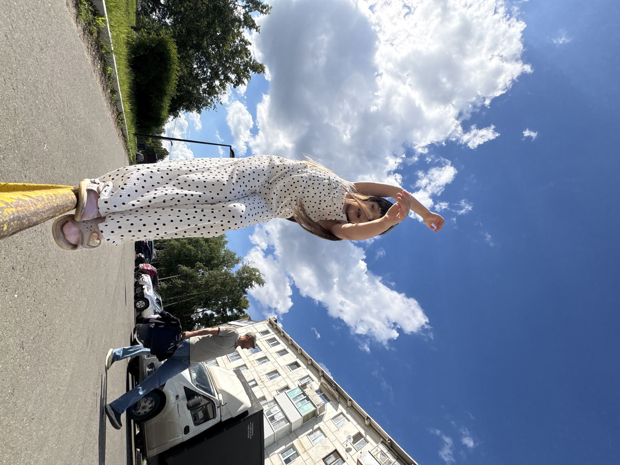 In Chaikovsky, Russia, a young girl in a white polka dot jumpsuit stands on a yellow railing, her arms raised in a playful gesture, against a backdrop of a bright blue sky with fluffy white clouds. To the right, a man in a grey t-shirt and blue jeans walks away from the camera, carrying a dark bag. Behind him, a white truck is parked next to a light-colored apartment building. The scene is set on a paved area, with green grass and trees visible in the background.