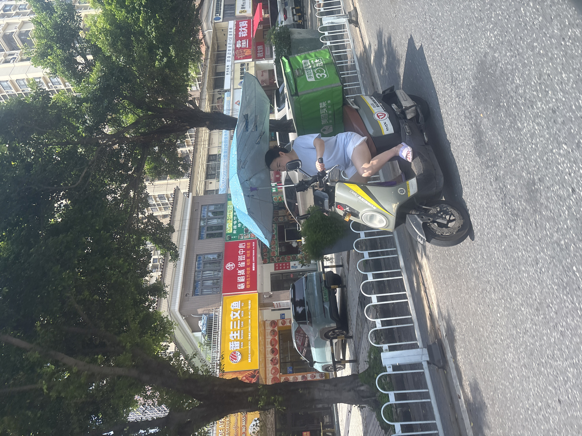In Baiyun District, China, a woman rides a scooter on a street. She is wearing a light blue t-shirt and white shorts, with a blue umbrella open above her to shield her from the sun. The scooter has a large green insulated bag strapped to the back, indicating she might be delivering food or goods. In the background, there are buildings with advertisements, a light-colored car parked on the side of the road, and trees providing shade. The street is paved and shows signs of traffic.