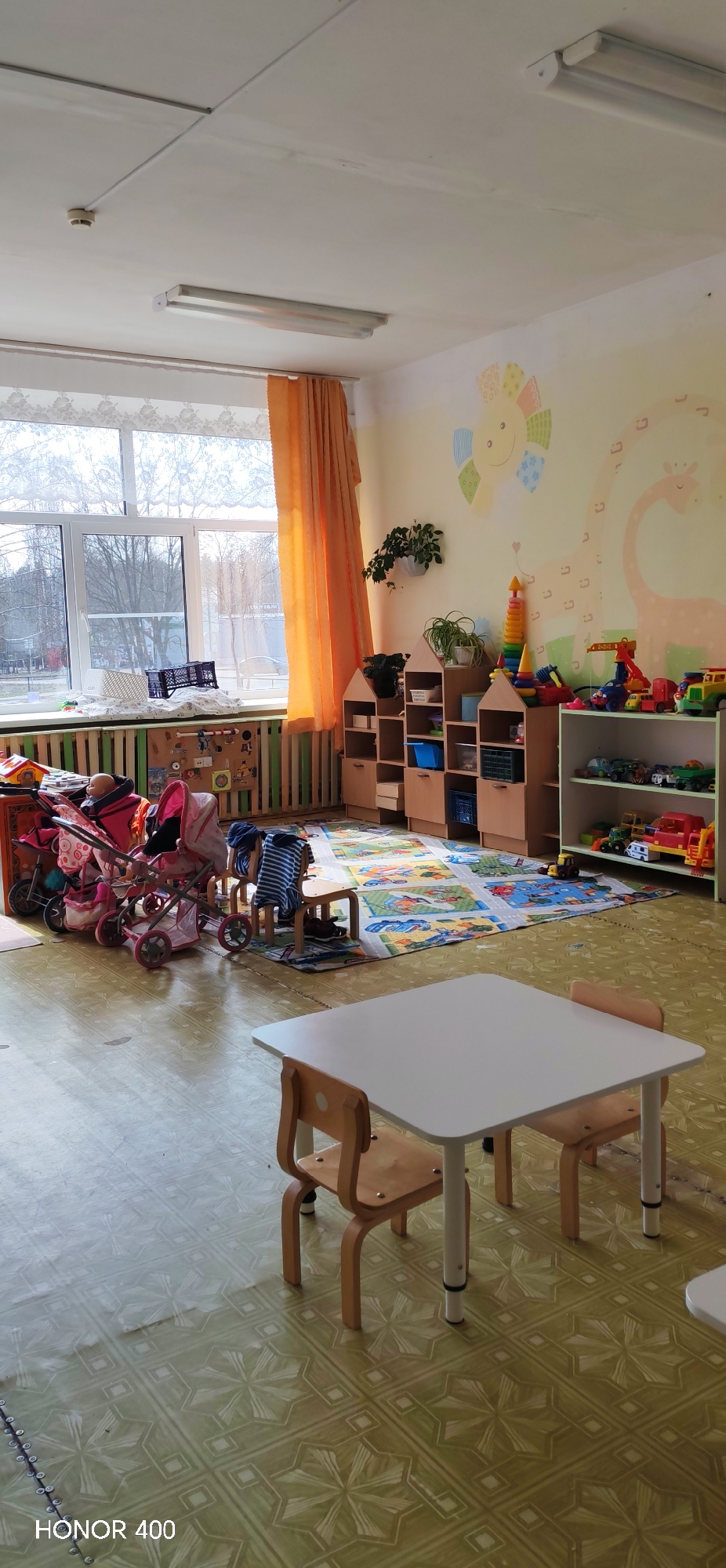 This image captures a brightly lit playroom or classroom in a children's facility, likely a kindergarten or daycare. No people are visible, but the room is fully equipped for children's activities.
The setting features light yellow walls adorned with colorful murals, including a sun-like figure and a large giraffe. Large windows covered by sheer white and orange curtains reveal a view of bare trees outside, suggesting a cooler season like late autumn or winter. Natural light streams into the room, complemented by fluorescent light fixtures on the white ceiling, which also has a visible smoke detector.
In the foreground, a small white square table is positioned with two small wooden chairs, indicating a children's activity area. The floor is covered with patterned linoleum in shades of green and yellow. Towards the middle ground, a colorful rug depicting roads and buildings lies on the floor, surrounded by various toys.
To the right, several shelving units are filled with toys and organizational bins. One unit is shaped like miniature houses. A white shelving unit holds numerous toy cars, trucks, and building blocks. Potted plants are placed on shelves and in a hanging planter on the wall. Near the window, a green slatted structure (possibly a radiator cover or cubby) is present, with baby doll strollers and a doll placed next to it. A small wooden stool and other small chairs are scattered around the play area.
The overall impression is one of a cheerful, organized space designed for young children's play and learning. A visible watermark in the bottom left corner of the image reads "HONOR 400".