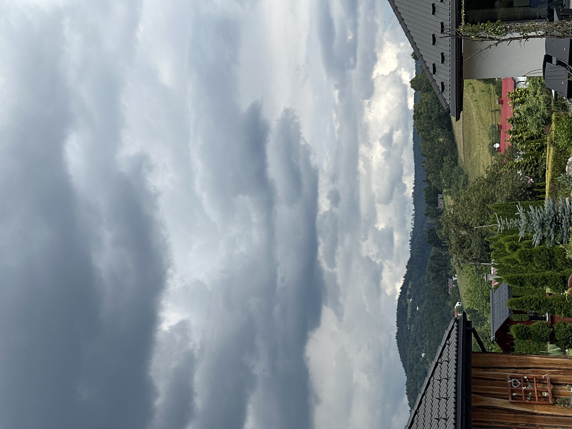 The image depicts a cloudy sky over a hilly landscape. In the foreground, two modern houses with dark tiled roofs are visible. The house on the left has a wooden facade and a small decorative window with a heart. The house on the right is white with a prominent roofline. Between the houses, various evergreen trees and shrubs are planted, creating a garden-like setting. In the middle ground and background, rolling hills covered in dense green forests rise up. Scattered among the trees are a few houses with red roofs, suggesting a residential area within the natural landscape. The sky is filled with dramatic, dark grey clouds, indicating overcast weather, possibly before or after a storm. The scene appears to be a rural or semi-rural residential area in the city of Wisła, Poland, characterized by its hilly terrain and lush vegetation under a moody sky.
