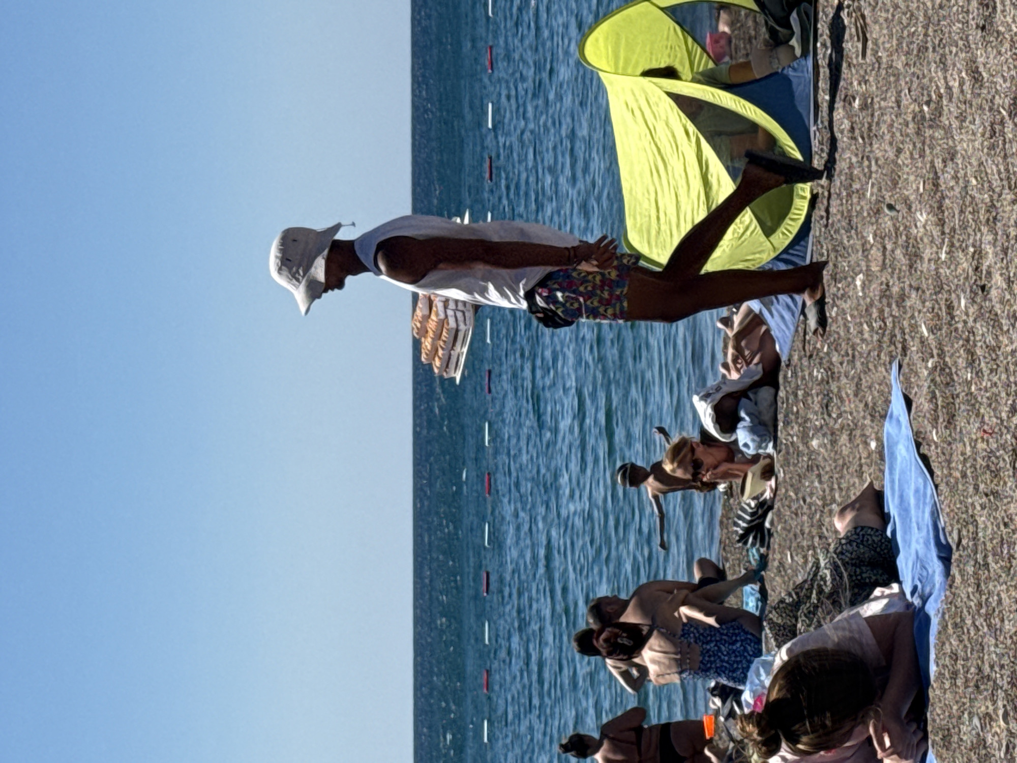 A vendor walks across a pebbled beach in Boreti, Montenegro, carrying a tray of pastries. The vendor, wearing a white bucket hat and a white sleeveless shirt, walks towards the right side of the frame. They are wearing patterned shorts and dark sandals. Several beachgoers are visible, some are seated on towels, others are in the water. A bright yellow and blue beach tent is set up on the right side of the image. The sea is calm and a clear blue sky is above.