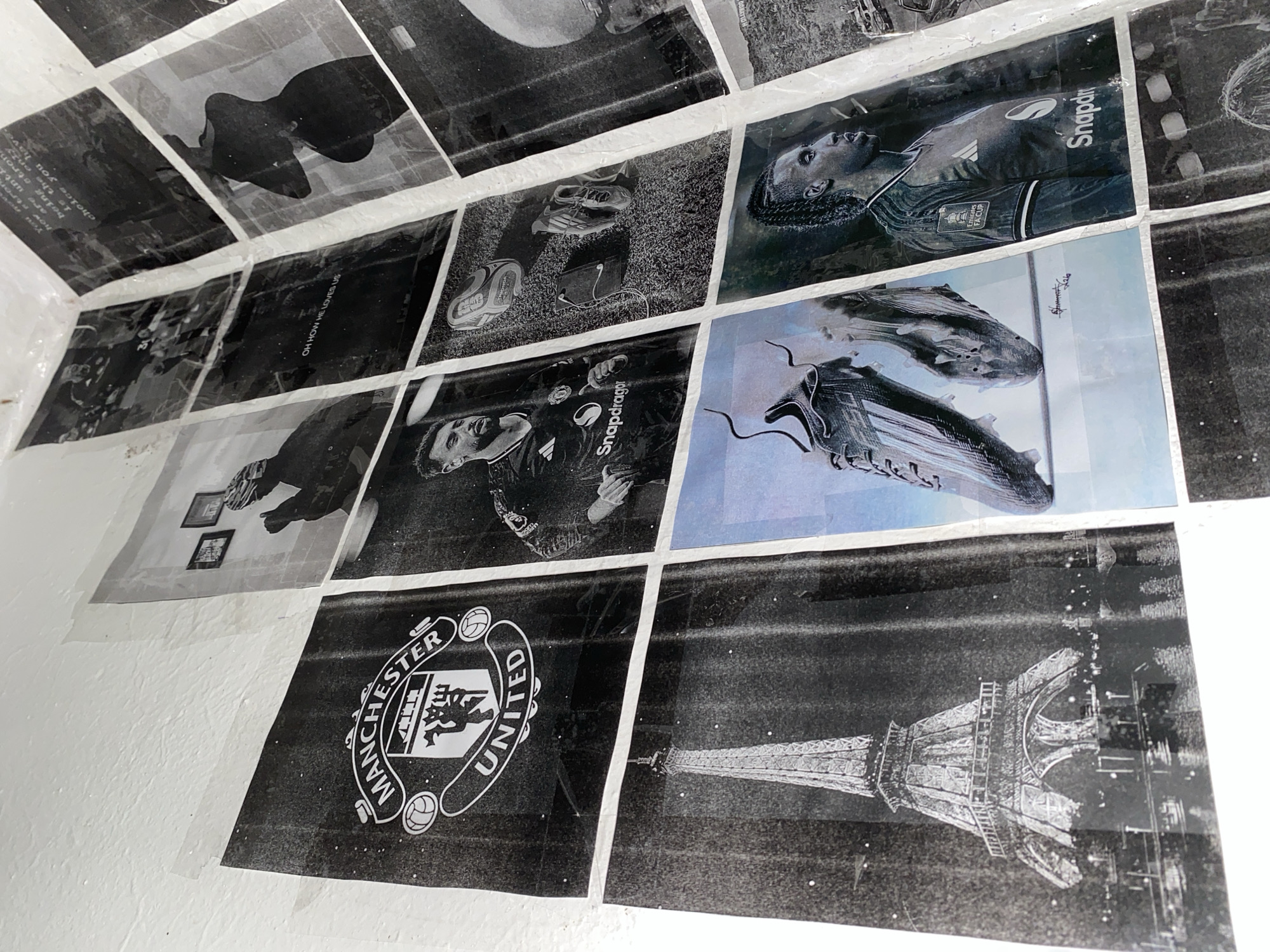 This media file is a collection of black and white photographs taped to a white wall, possibly in a bedroom or living space. The photographs are arranged in a grid-like fashion, overlapping in some areas.
The subjects in the photographs are predominantly related to football, featuring images of soccer players in action, football boots, and sports equipment. There are also close-ups of players' faces and dynamic shots of them on a grass field. Some images show text, including sponsor logos like "Snapdragon" and "Emirates FA Cup," and what appears to be motivational or team-related slogans. One photograph features a silhouette of a person, while another displays the Manchester United Football Club logo. In the lower right corner, an image of the Eiffel Tower is visible. The overall impression is that of a fan's tribute or a personal collection of football memorabilia. The lighting in the photographs varies, with some appearing to be taken outdoors during the day and others in a more controlled studio setting. The presence of these images on a wall suggests a display intended to be viewed and appreciated.