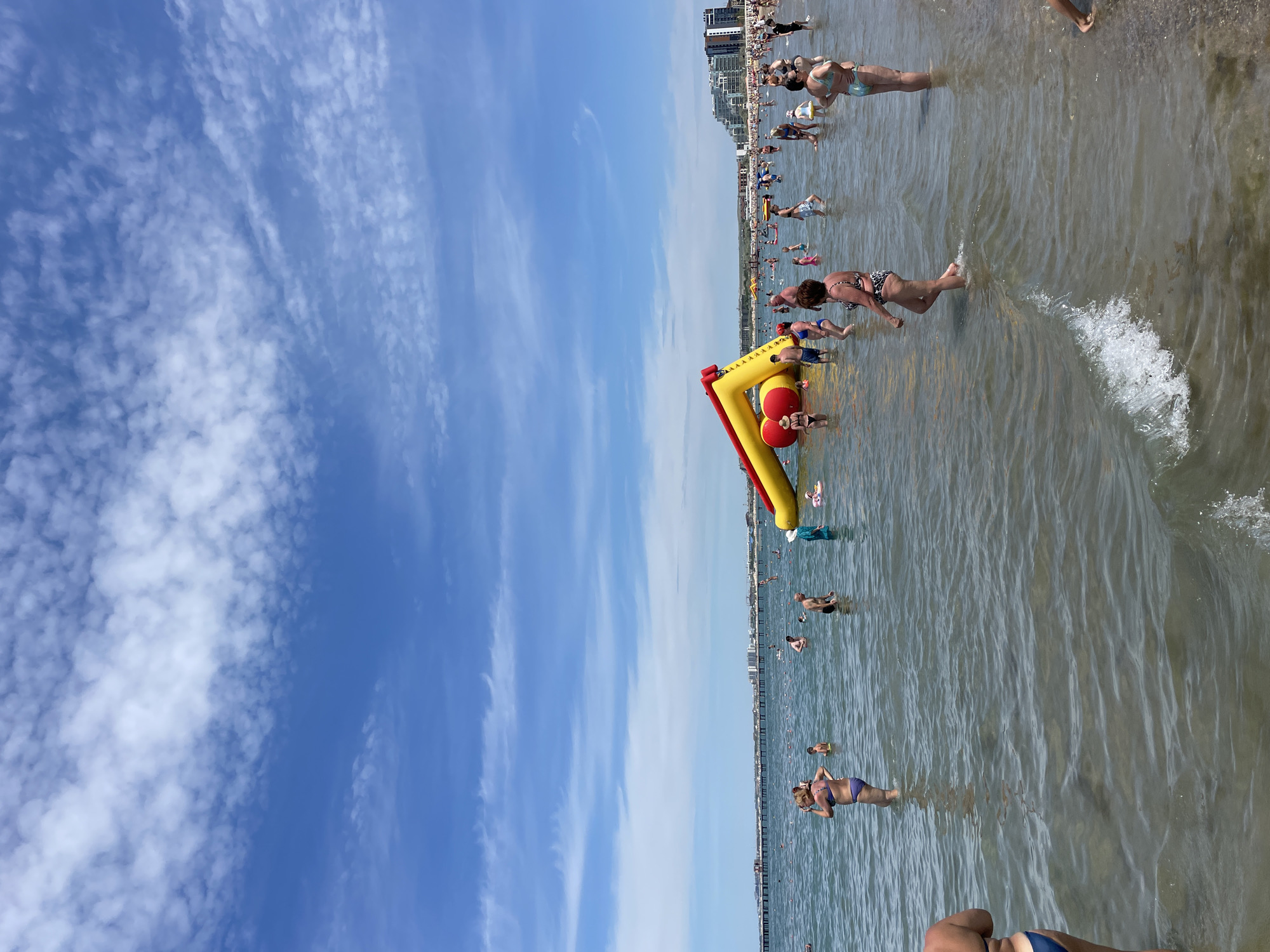An aerial shot shows a crowded beach on a sunny day with scattered clouds. The water is clear and shallow, with many people swimming and wading. An inflatable water slide is set up in the water, and children are playing on it. A pier extends out into the water in the distance, and buildings line the shore.