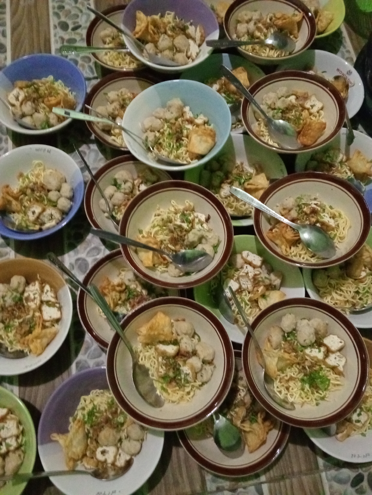 An overhead medium shot captures a close-up of multiple bowls of noodles and meatballs, seemingly prepared for a meal in Krajan Kidul, Indonesia. The food is arranged on a tiled surface that is a mosaic of earthy tones. The bowls themselves are varied, some with brown rims, others white, and one with a blue hue, all containing a generous portion of noodles topped with meatballs, fried dumplings, and chopped green onions. Spoons are placed in several of the bowls, suggesting an imminent meal. The lighting appears to be dim, likely from an interior source, casting subtle shadows and highlighting the textures of the food. The arrangement implies a gathering or a communal meal.