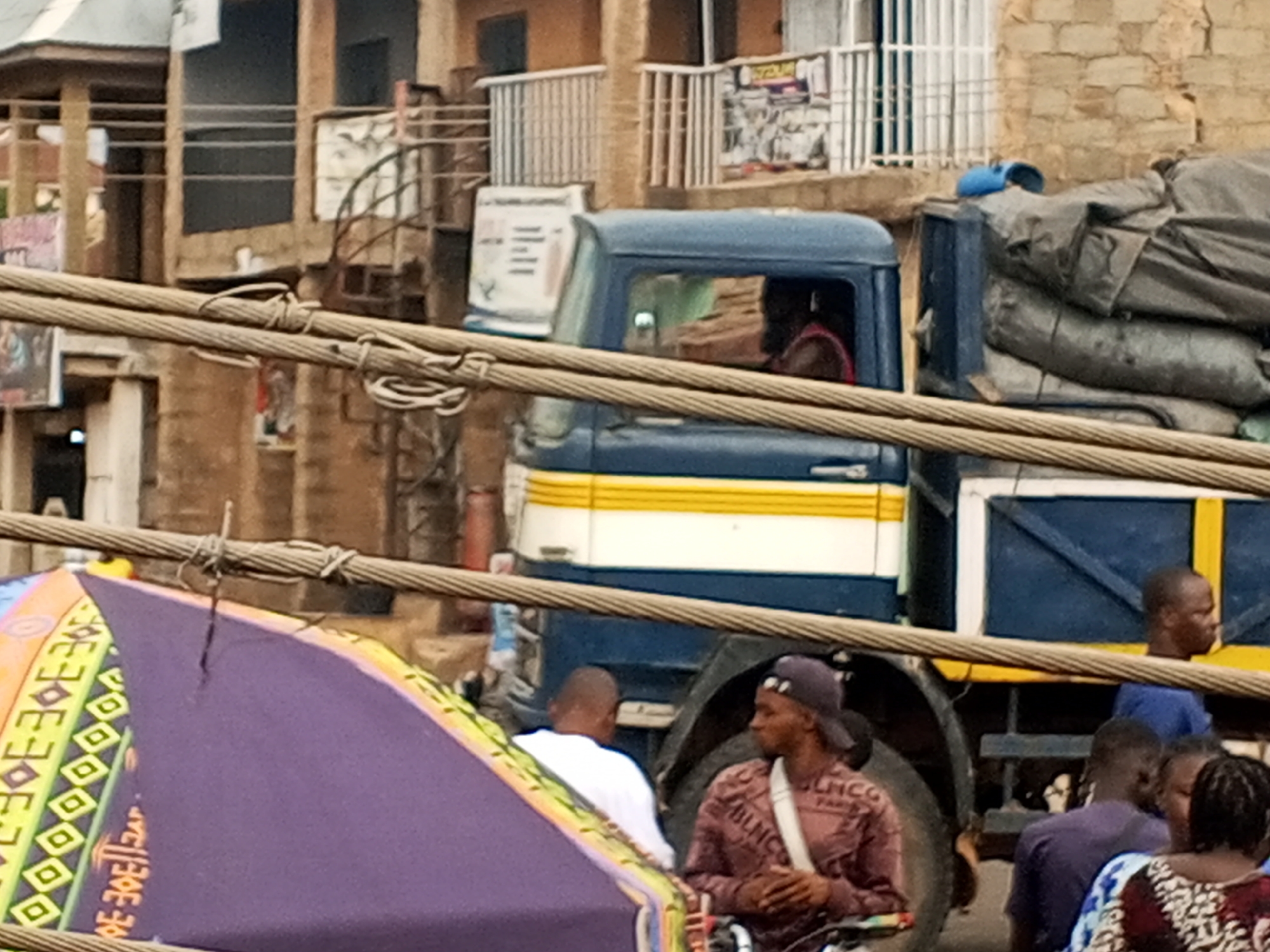 The image shows a street scene in Gwong, Nigeria. A blue and white truck, loaded with bundled items covered in a gray tarp, dominates the right side of the frame. The truck's cabin is visible, with a driver inside. Several people are gathered around the truck, including a man in a white shirt, a man wearing a purple baseball cap and a brown shirt with a white strap, and a man in a blue shirt. A partially visible purple and orange tent-like structure is on the left, with an elaborate pattern. Overhead, thick power lines run across the foreground, slightly blurring the background. In the background are several buildings, with one appearing to have metal staircase. The scene appears busy and the lighting is slightly overcast.