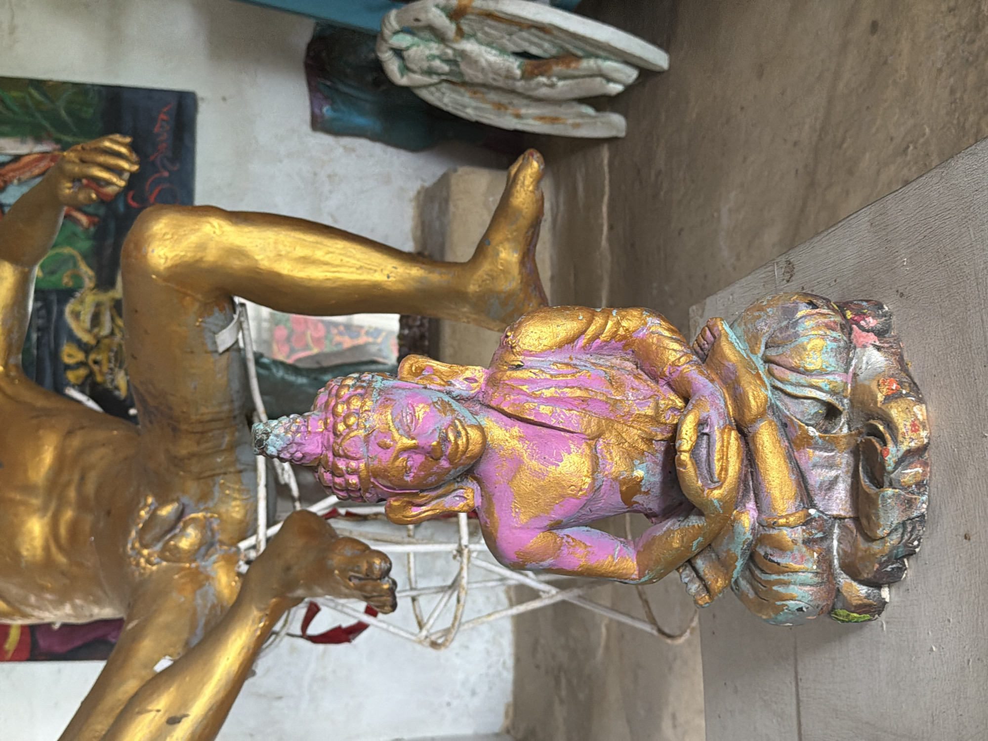 In a dimly lit room, a small, colorful statue of Buddha sits in the foreground. The statue is made of what appears to be resin or plaster, painted with shades of pink, gold, and turquoise, depicting the Buddha in a meditative pose. Behind the Buddha, a large, golden statue of a human figure is partially visible. This figure is in a seated position, with its legs spread and one foot resting on a white metal stand. The golden statue has a metallic sheen and appears to be made of a solid material like bronze or fiberglass.
To the right of the golden statue, a white, angel-like figurine is positioned. It has wings and appears to be made of wood or a similar material, with some areas of paint chipping away. The background of the scene shows a textured wall and hints of other art pieces, including a painting with abstract shapes. The floor is a mix of concrete and what seems to be wooden planks. The overall impression is that of an artist's studio or a place where art is displayed, potentially in Tejakula, Indonesia, given the mention of the location. The lighting is subdued, casting soft shadows and highlighting the textures and colors of the various objects.
