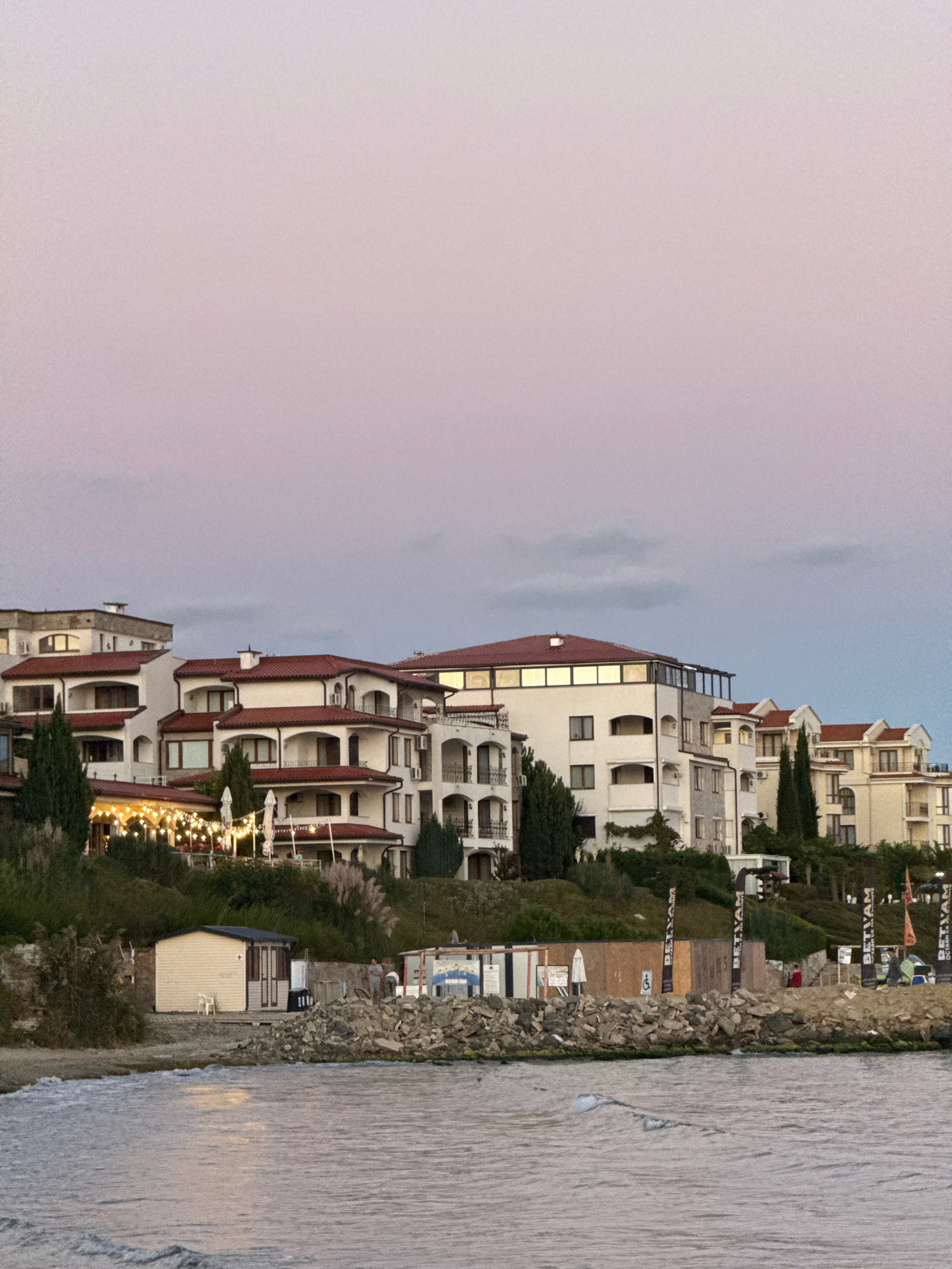 The image shows a coastal scene in Sveti Vlas, Bulgaria, at dusk. The sky is a gradient of soft pink and pale blue. Dominating the upper half of the image are several multi-story buildings with white facades and red tiled roofs, situated on a hillside. Some of these buildings feature balconies and large windows.
Below the buildings, a green, overgrown hillside slopes down towards the water. There are trees and bushes, including tall cypress-like trees and some smaller shrubs. String lights are visible on some of the buildings' terraces, suggesting an outdoor dining or seating area.
In the foreground, a rocky shoreline separates the sea from the land. On the shore, there are several small structures, including a light-colored, shed-like building with a dark roof and door, and a larger wooden structure with various signs and posters attached. One sign clearly displays the word "BETVAM" multiple times. Beach umbrellas and what appear to be beach chairs or loungers are visible near these structures.
The sea is calm with gentle ripples on the surface, reflecting the subdued light of the sky. A few dark rocks are submerged in the water near the shore. The overall impression is one of a tranquil evening by the sea in a resort town.