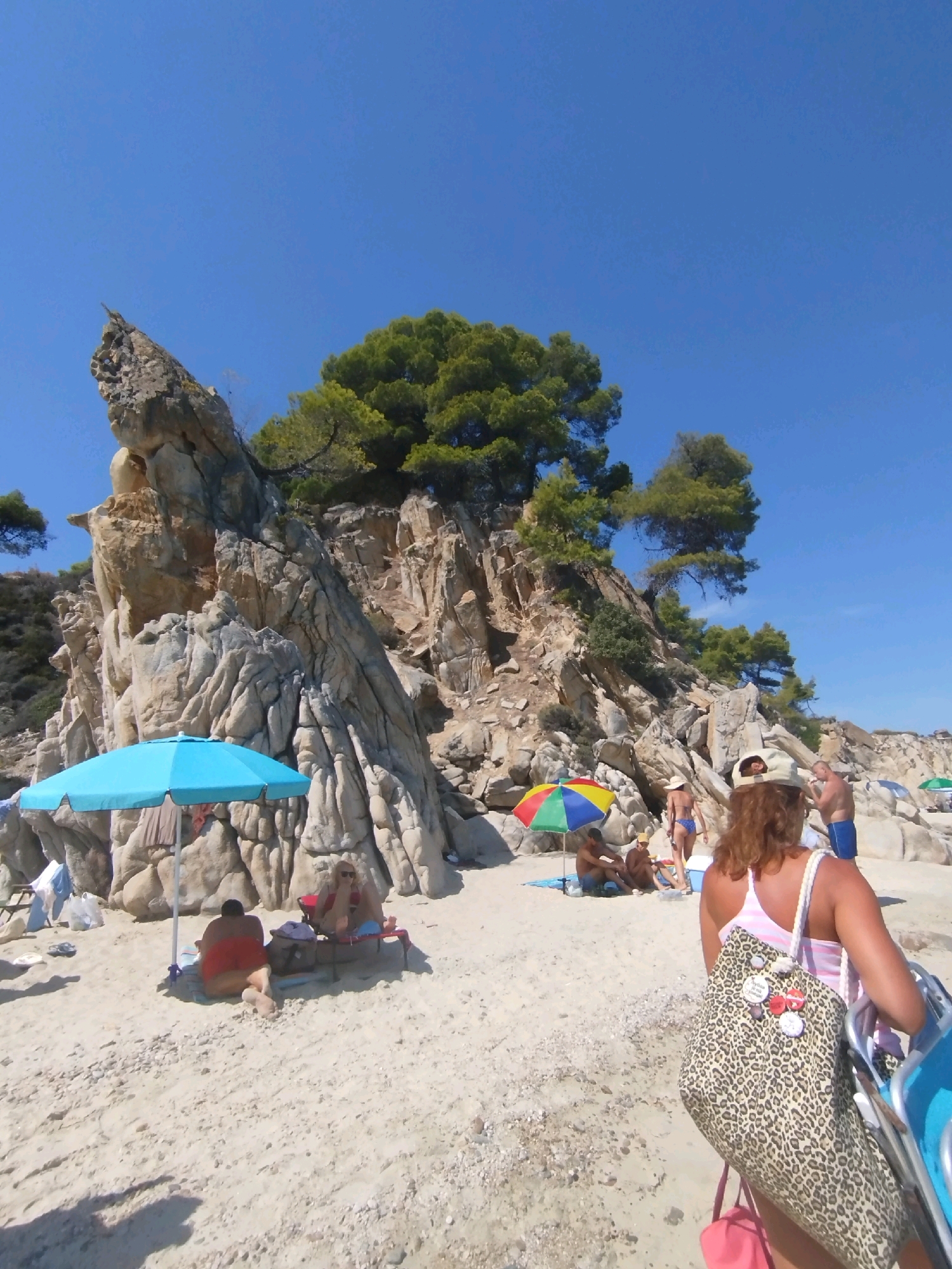 The image depicts a sunny day at a beach in Sithonia, Greece, with a rocky cliff face in the background and several people enjoying the seaside. A large, tan rock formation with trees growing from its top dominates the left side of the frame. In the foreground, a woman with reddish-brown hair and a tan hat is walking away from the camera, carrying a large leopard-print bag. She is wearing a pink and white striped tank top. To her right, a man in blue shorts is visible. On the sandy beach, various people are relaxing under umbrellas. A bright blue umbrella shades two individuals sitting on chairs, one in red shorts and another whose face is obscured by a hat and sunglasses. Further back, a colorful umbrella shelters a group of people, and another green umbrella is visible in the distance. The sand is a light tan color with scattered small stones and pebbles. The sky is a clear, bright blue.