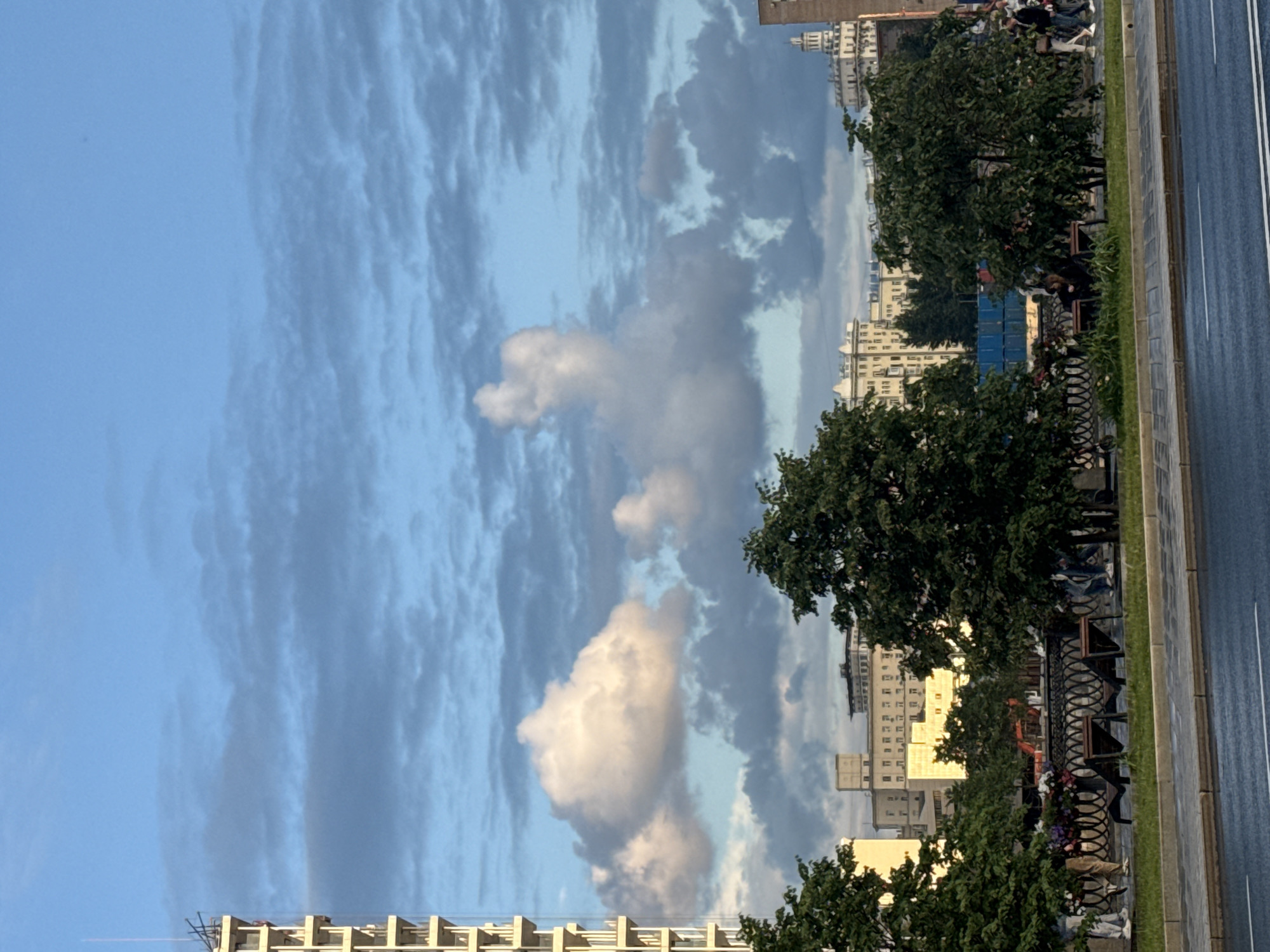 The image depicts a street scene in Moscow, Russia, with a focus on the sky and cityscape. A tall, light-colored building with a modern, angular design stands prominently on the left side of the frame, its upper section reaching into the sky. The sky itself is a vibrant blue, interspersed with dramatic, textured clouds that create shadows and highlights across the scene.
In the mid-ground, several trees with dense green foliage are visible, some partially obscuring the buildings behind them. The buildings in the background are varied in architecture, suggesting a mix of older and newer structures typical of a large city. One building has a white, domed roof, adding a distinct visual element to the skyline.
In the foreground, a wide asphalt road with lane markings runs horizontally across the bottom of the image. A paved sidewalk with a grassy verge borders the road, and a decorative metal fence runs along the edge of this area. Several people are walking along the sidewalk, their figures small in comparison to the urban landscape. Two empty lounge chairs are positioned on the grassy verge in front of the fence, facing away from the camera. A few dark-colored benches are also visible along the walkway. The overall impression is one of a bustling city on a day with dynamic cloud formations.