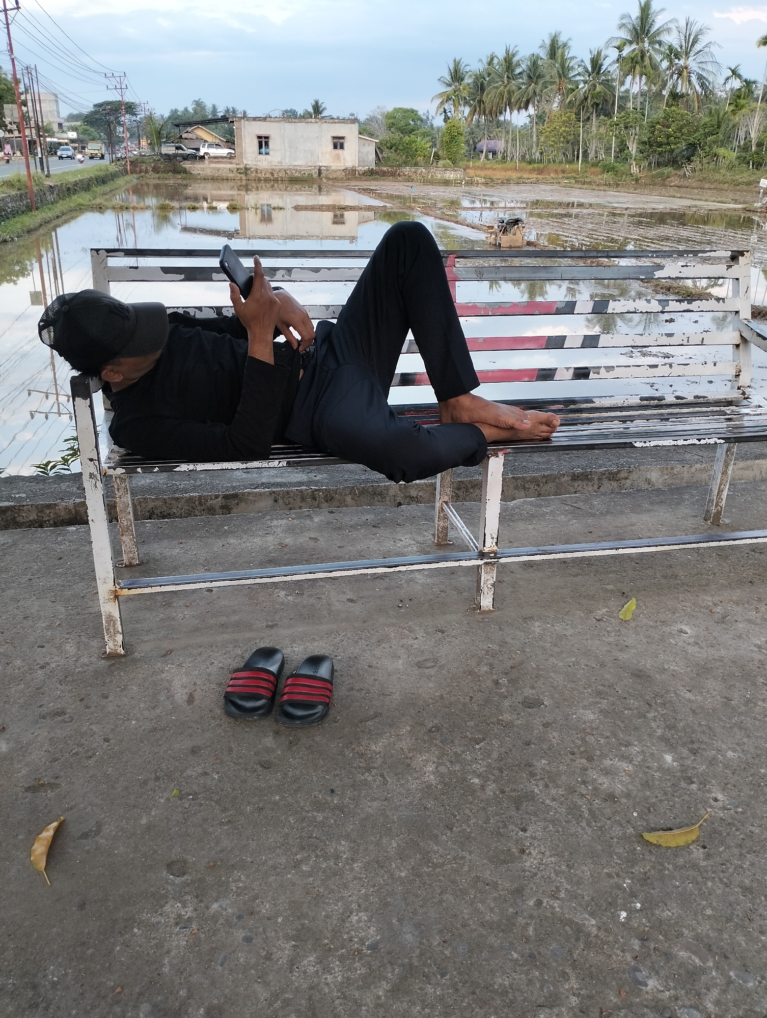 A person wearing black clothing and a black baseball cap lies on their back on a weathered white and red striped bench. They are holding a black smartphone in their hands, appearing to be looking at it. The bench is situated on a concrete surface next to a body of water that reflects the sky and surrounding environment. In the background, there is a flooded rice paddy with rows of vegetation, coconut trees, and a few buildings. A road with vehicles is visible in the distance to the left. A pair of black and red striped flip-flops rests on the concrete near the bench. The sky is overcast with a hint of blue. The location is identified as Blangmetimu, Indonesia.