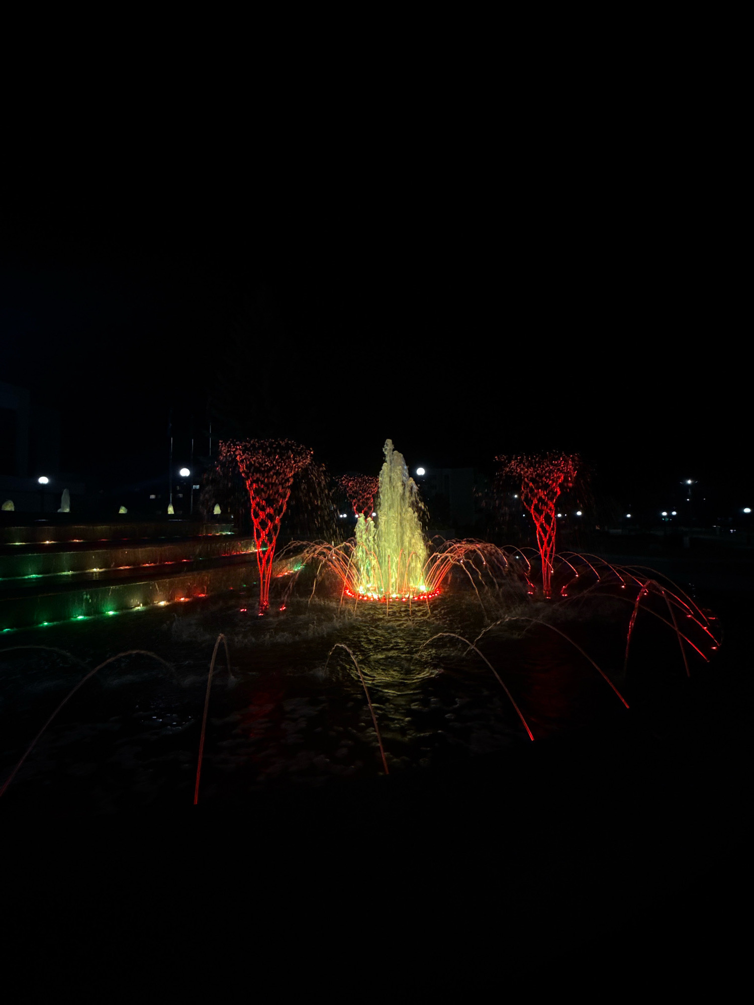 The image depicts a public fountain illuminated with colored lights at night. The fountain features multiple jets of water, some of which are lit with bright yellow and red hues. The water cascades down tiered steps, also lit with green and red lights, creating a vibrant visual display against the dark background. In the distance, subtle lights from buildings and streetlamps are visible, suggesting an urban environment. The scene is set in Mikashevichy, Belarus, a city known for its industrial and natural landscapes, and this fountain likely serves as a recreational focal point. There are no people visible in the frame, and the focus is solely on the illuminated water feature and its surroundings.