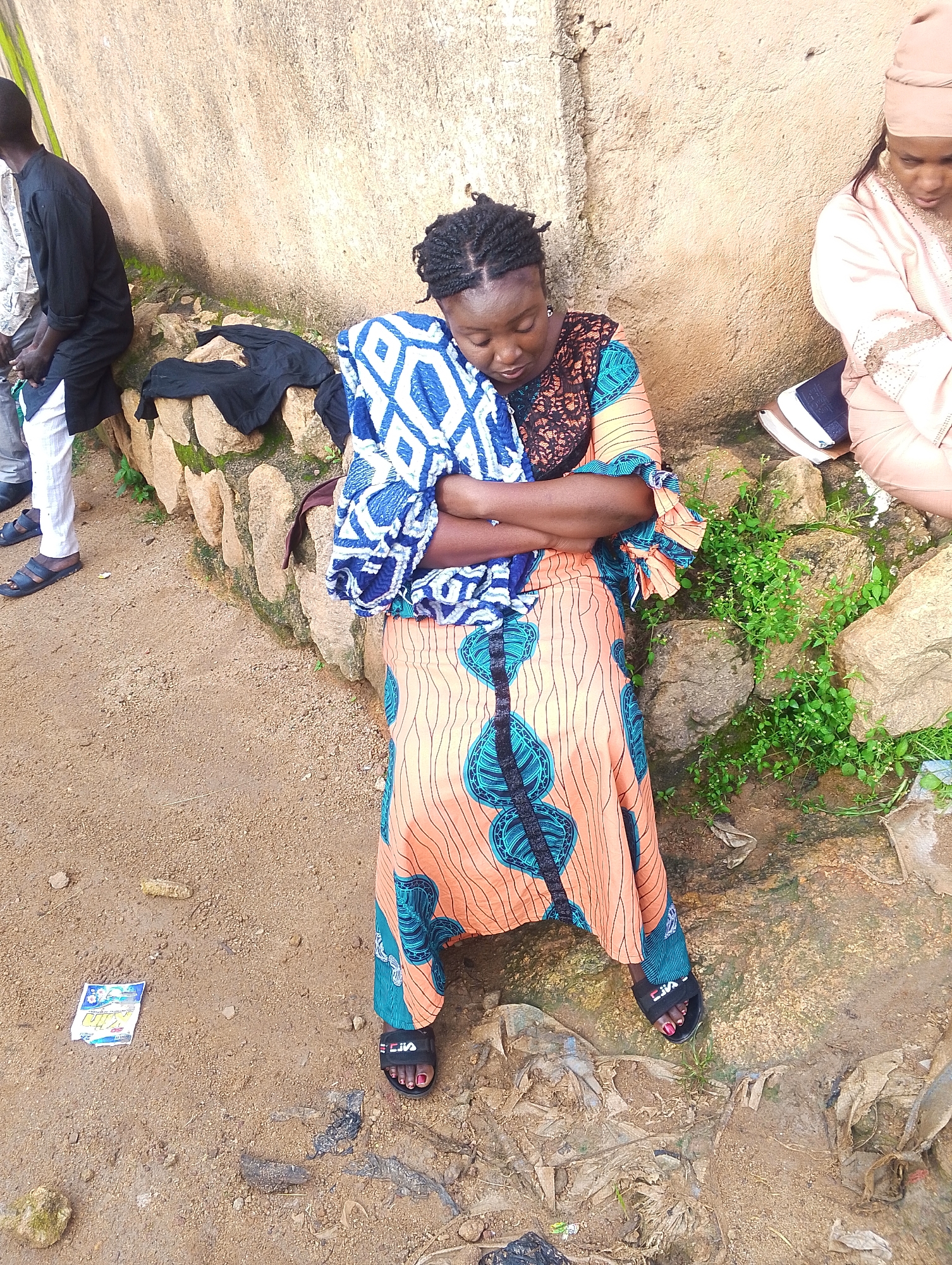 An overhead, full shot shows a woman with dark skin and braided hair sleeping. She is wearing a vibrantly patterned orange dress with teal and black designs. She is holding a blue and white patterned blanket to her chest. Her arms are crossed over the blanket, and her head is bowed. She is wearing black sandals with a white logo. She is sitting on a stone wall. To her left, a person in a black shirt and white pants is visible. To her right, another person in a pale pink outfit is sitting. The environment appears to be outdoors with a rough, dirt ground and a textured, tan-colored wall behind the subjects. There are rocks and some green vegetation around the stone wall.