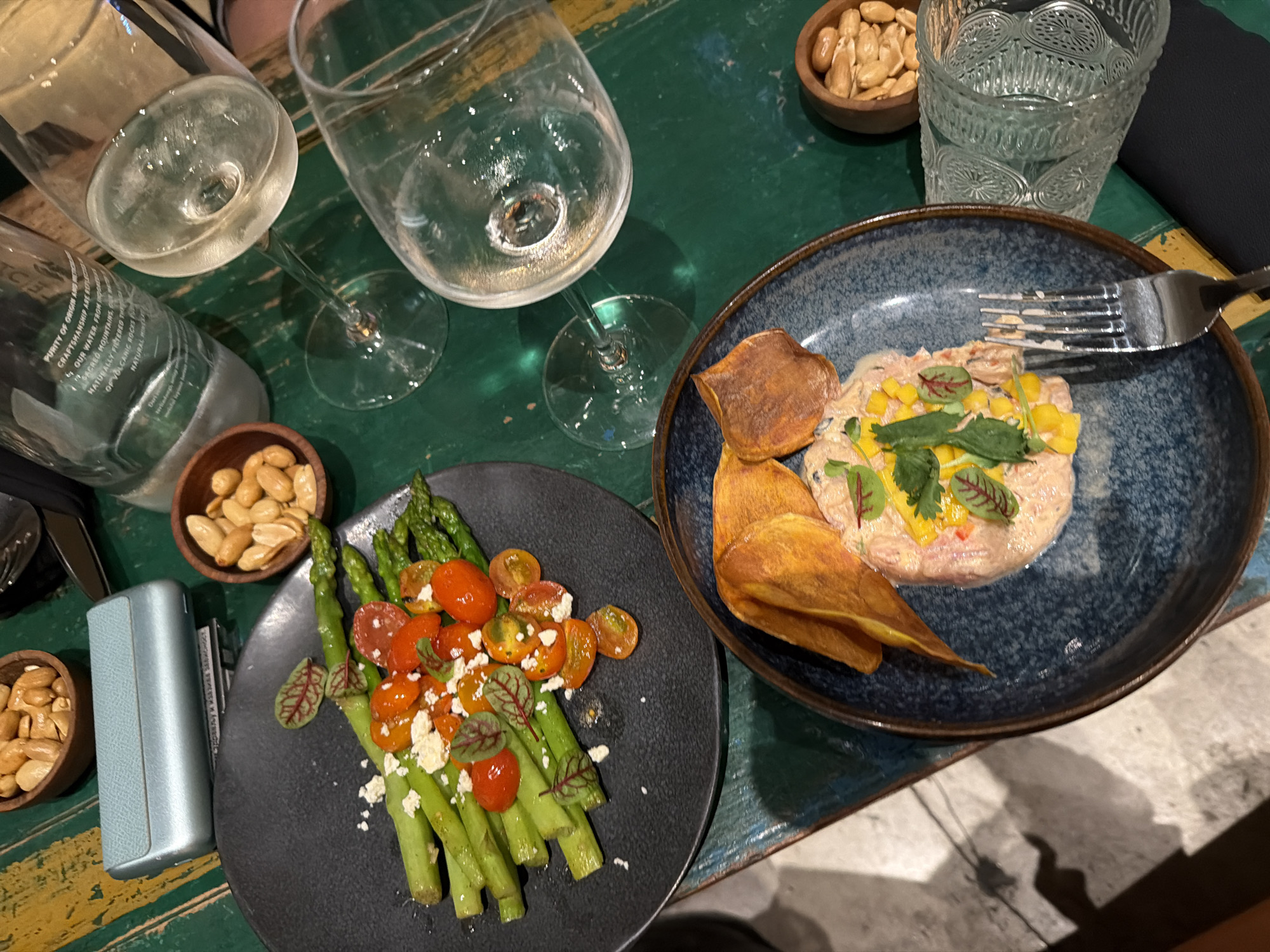 The image captures a close-up, overhead view of a dining table set with two plates of food, wine glasses, and small bowls of nuts. The table itself is a rustic, green wooden surface with visible wear.
On the left, a dark plate holds a dish of asparagus spears topped with halved cherry tomatoes and crumbled white cheese, garnished with small red-veined greens. To the right, a blue-rimmed dark blue bowl contains a dish that appears to be a seafood tartare or ceviche, mixed with diced yellow and red ingredients, garnished with fresh herbs. Two crispy, golden-brown chips, likely sweet potato, are placed on the side of this dish.
Two wine glasses, one partially filled with white wine, are visible. A clear glass with a decorative pattern sits behind the food. Two small wooden bowls filled with nuts, possibly almonds, are also present on the table. A silver object, possibly a cigarette lighter or portable battery, lies next to the left plate. A partially visible bottle with text printed on it is also in the frame.
The lighting suggests it is daytime, possibly late afternoon or early evening, given the ambient light and shadows. The scene evokes a casual dining experience. The visible text on the bottle fragment includes "TCEL," "PURITY OF ORIGIN AND," "OUR WATER FROM INDONESIA," and "SACRED MOUNTAINS." The location context is North Kuta, Indonesia.