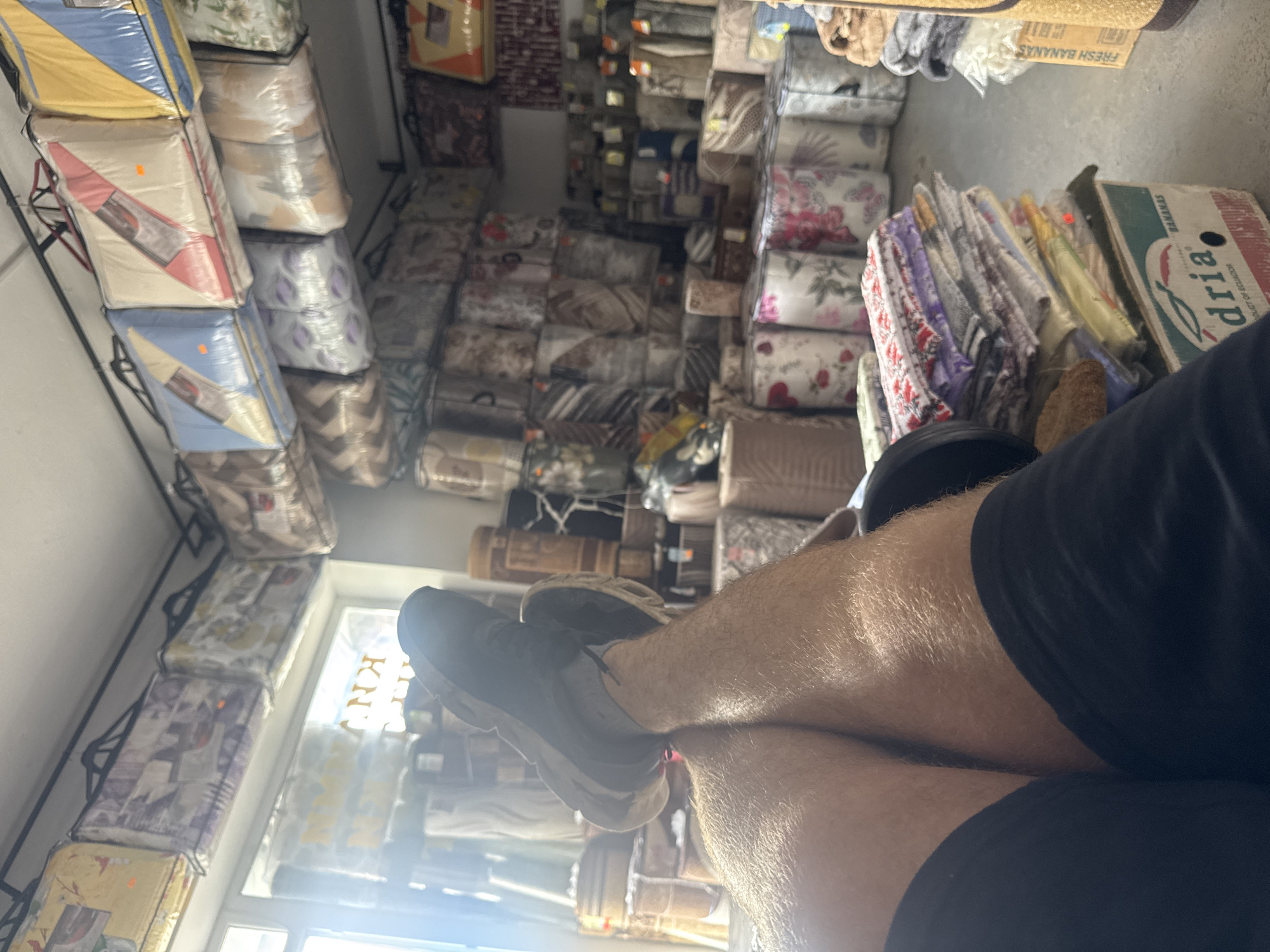 The image depicts the interior of a retail establishment in Shumen, Bulgaria, specializing in bedding and textiles. The foreground features a person's legs and feet, casually crossed, with one foot resting on the other. The individual is wearing dark athletic shoes and black shorts, with visible leg hair.
The background is filled with merchandise, predominantly bedding sets packaged in clear plastic bags. These are neatly arranged on shelves and racks, extending throughout the visible space. Various patterns and colors are apparent on the packaging, suggesting a wide selection of products. Among the textile items, some appear to be rolled fabric or carpets. A cardboard box labeled "BANANAS" with the brand name "Adria" is visible in the lower right quadrant, suggesting a possible secondary use of the space or adjacent area for storage of diverse goods. The overall lighting suggests an indoor setting, possibly with natural light filtering in from a window, hinted at by a bright, slightly out-of-focus area on the left. The store appears to be well-stocked.