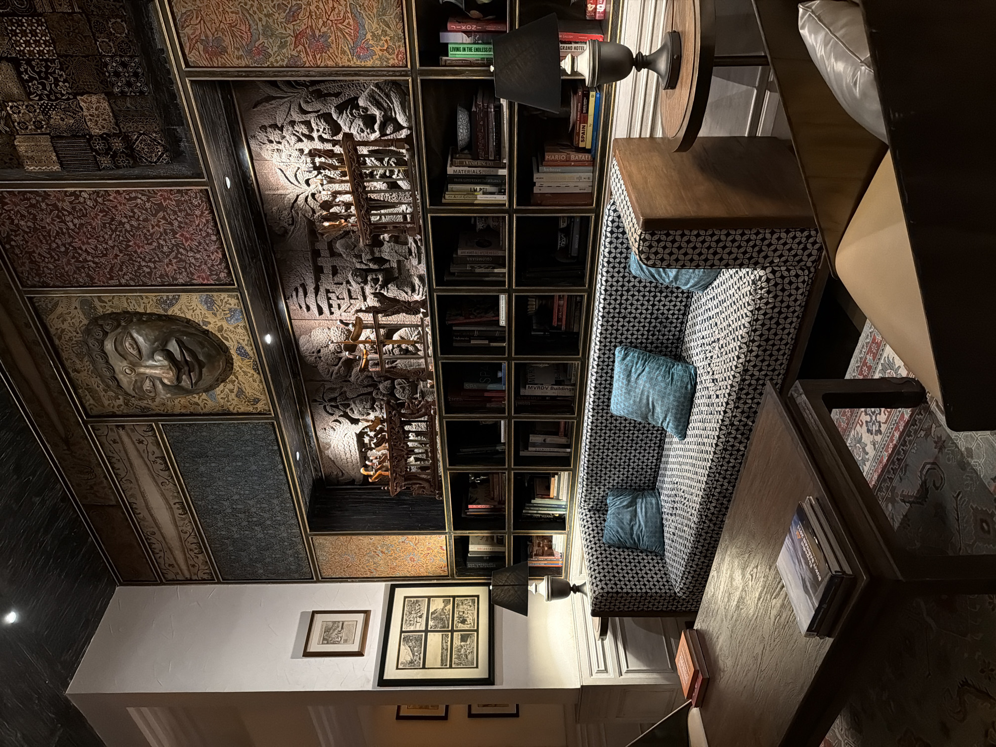 The image shows the interior of the Jikoni restaurant. The restaurant has a unique design with various patterns and textures on the walls and ceiling. There is a seating area with a patterned sofa, blue cushions, and a wooden coffee table. The room also features a bookshelf with various books and decorative items. The exact location in the city of Unknown, Unknown cannot be determined from the image.