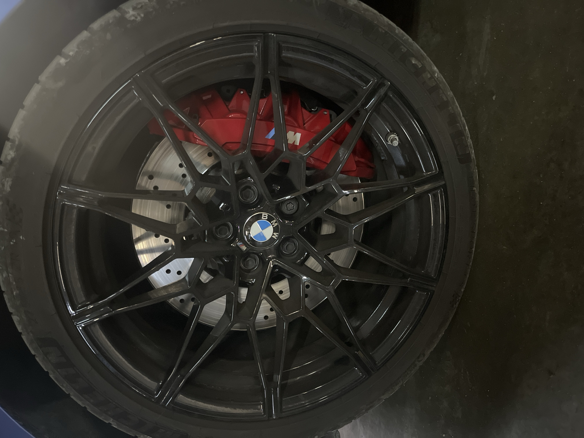 The image shows a close-up of a BMW wheel, specifically a black multi-spoke alloy wheel with a BMW logo at the center. Behind the wheel, a perforated brake disc and red brake calipers with an "M" logo are visible. The tire is black and appears to be slightly worn. The wheel is on a dark, presumably paved surface. There is no indication of the location being Varna, Bulgaria, or any other specific city. No people, actions, or interactions are visible in the image. The environment is undefined beyond the immediate vicinity of the wheel and tire.