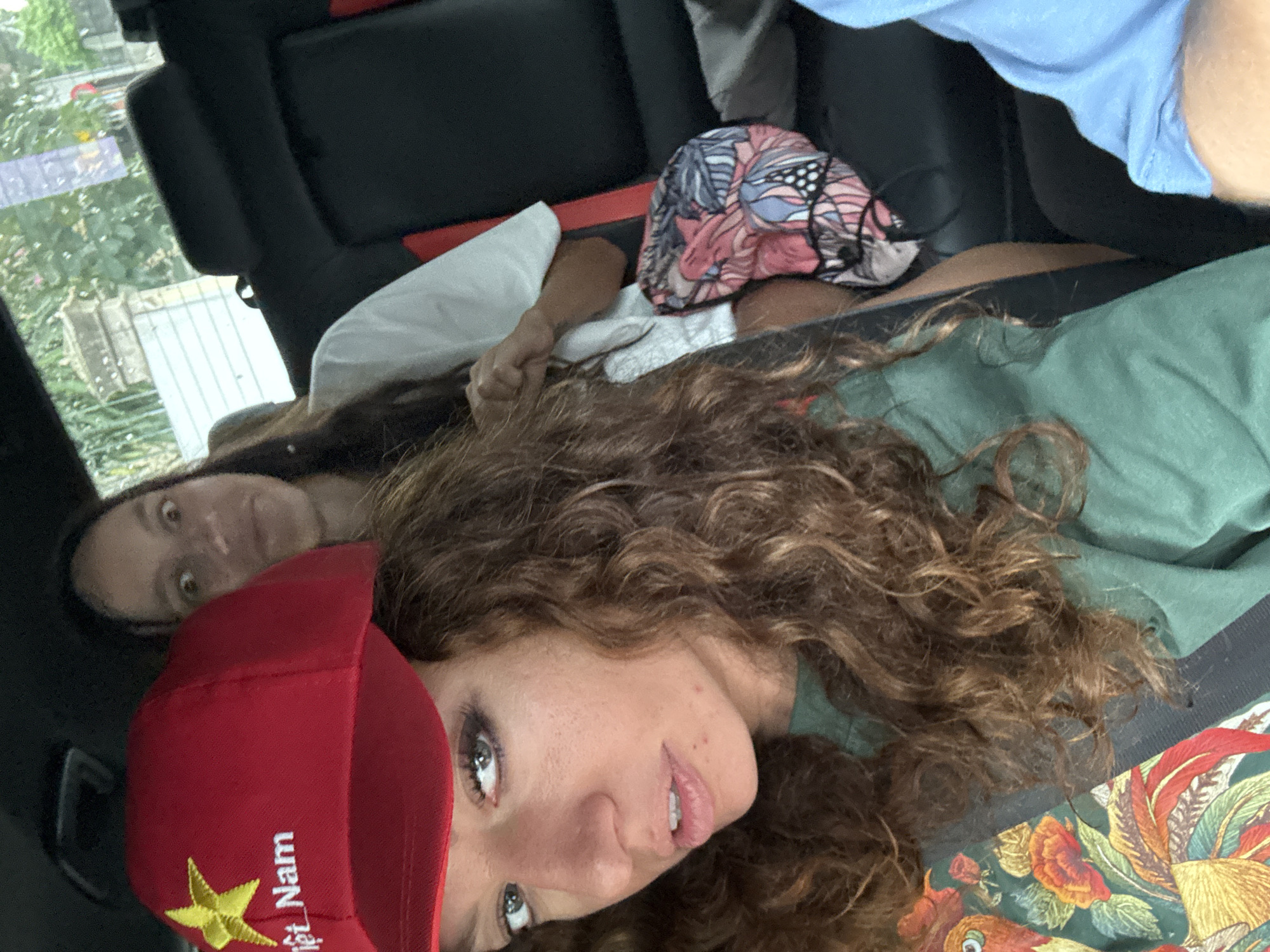 Two women are in the back seat of a vehicle. One woman wears a red hat with "Việt Nam" and a gold star emblem. The background visible through the car window suggests an outdoor location, and the hat indicates a possible association with Vietnam, though Nghia Lo Ward is not explicitly discernible. The car's interior is dark, with seats and upholstery visible. One woman's hand is visible. The exact interactions and actions are not clear, but they are positioned closely together in the vehicle.