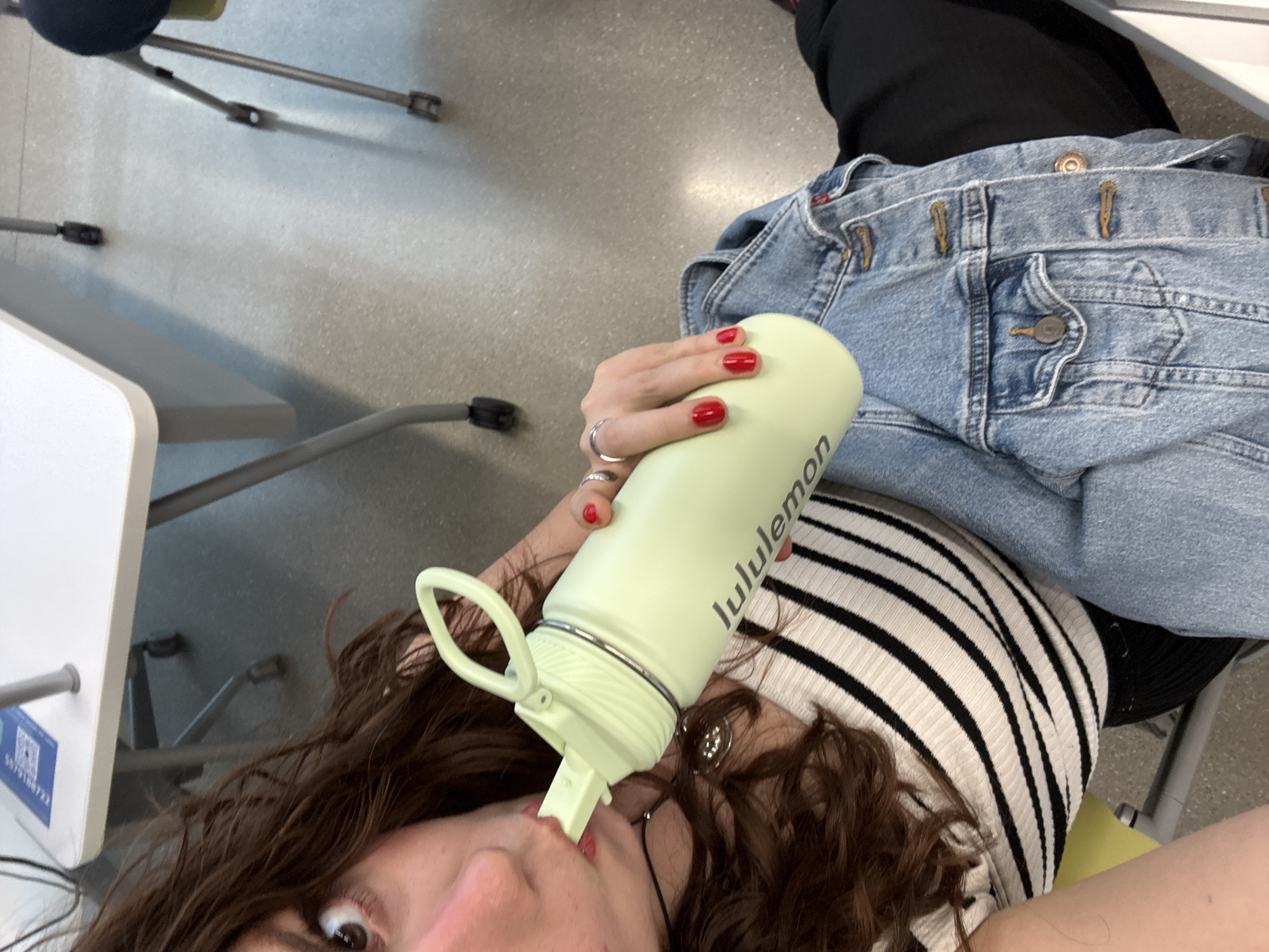 This rotated close-up image captures a person drinking from a light green Lululemon water bottle in an indoor setting. The central figure has medium-brown, wavy hair, with one wide-open brown eye visible, looking upwards. Their mouth is engaged with the straw of the water bottle.
The person's hand, adorned with several silver rings and painted with bright red nail polish, holds the bottle. They are dressed in a black and white striped long-sleeved top, partially obscured by light-wash denim overalls or jeans visible across their lap.
The background shows a smooth, light gray floor and the metallic legs of what appear to be chairs or tables, suggesting a functional space like a classroom, library, or office. To the left, a white desk or table surface is visible, bearing a sticker with a QR code and the numerical sequence "5079188722". The lighting is consistent with an indoor environment, providing no specific cues for the time of day. The visible text "lululemon" is prominently displayed on the water bottle. The scene depicts a casual, everyday moment of hydration.