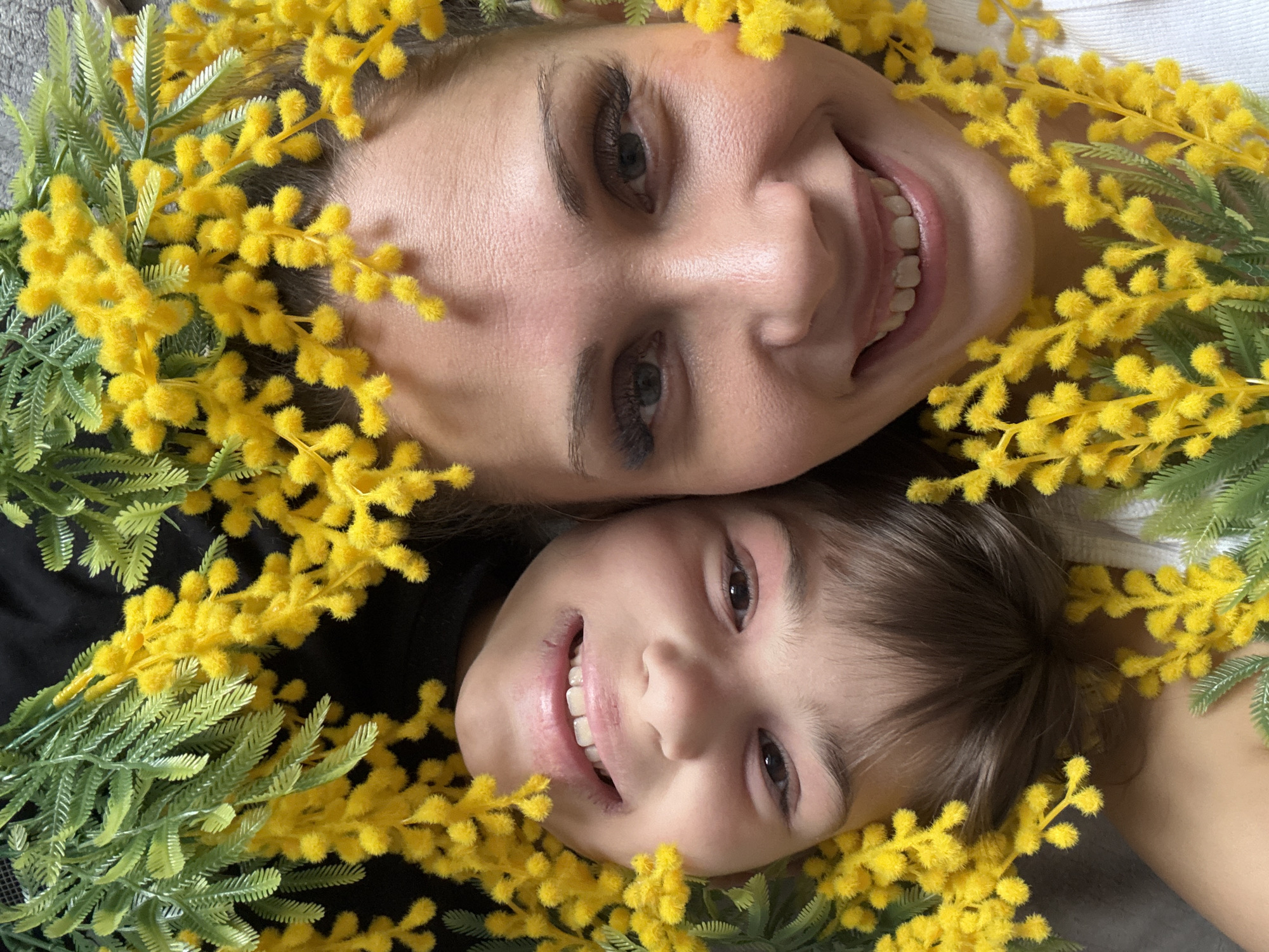 A close-up, overhead shot features two smiling faces surrounded by yellow flowers with green leaves, possibly mimosa. The faces appear to be those of an adult female and a child, possibly a girl, positioned close together. The background is blurred.
The faces appear to be illuminated from overhead lighting, and their complexions are clearly visible.
There is no indication of the location in the city of Rusanovo, Russia, or any other location.