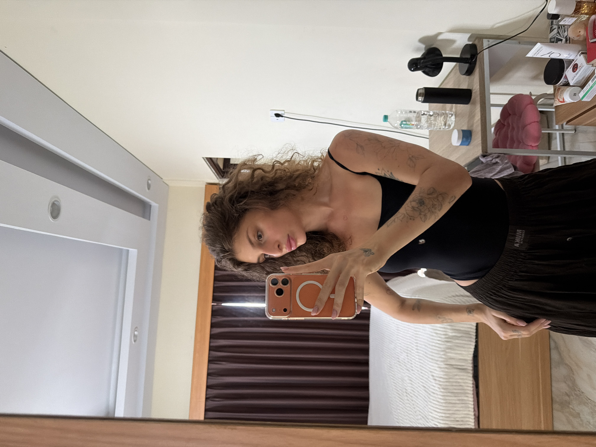 The image depicts a woman taking a selfie in a bedroom. She is standing in front of a mirror, holding an orange smartphone. She is wearing a black tank top and a dark brown skirt. The room appears to be a hotel room, with a bed visible in the background, along with a table with various items on it, including a lamp, a water bottle, and some toiletries. The walls are a neutral color, and there are curtains behind the bed. The ceiling has recessed lighting. The scene is consistent with a typical hotel room in North Kuta, Indonesia, a popular tourist destination known for its beaches and resorts.