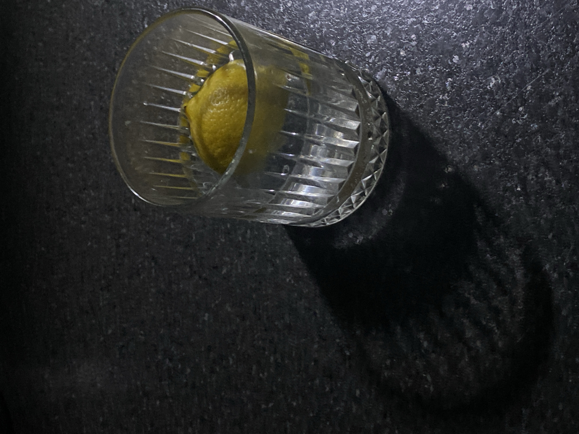 A lemon is in a cut glass tumbler on a dark surface. The light source casts a shadow to the right. The scene is undifferentiated. There are no people present. No location information is discernible.